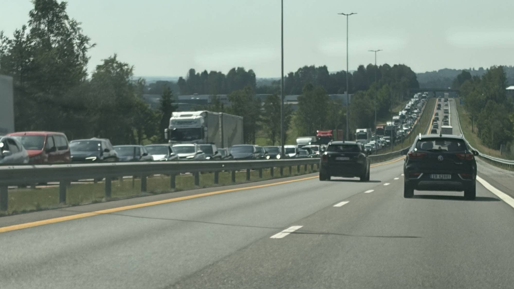 Kø på motorvei med mange biler og lastebiler i begge kjøreretninger under åpen himmel. (Bildebeskrivelsen er laget av en KI-tjeneste)