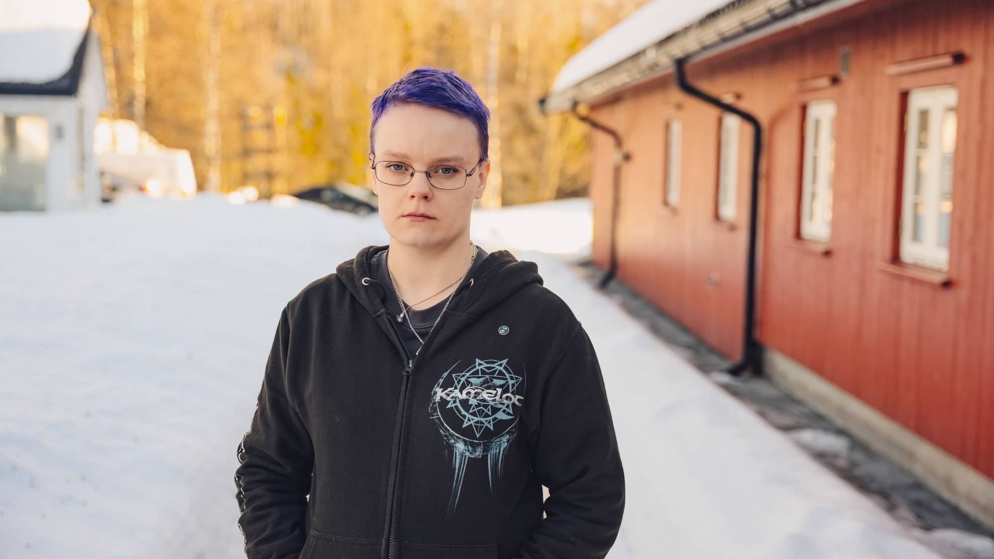 En dame med briller, lilla hår og svart hettegenser står ute i snøen ved et rødt trehus.