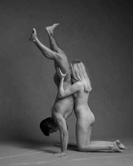 En mann med mørkt hår og tatoveringer står på hendene. En blond kvinne kneler bak ham og holder beina hans. Ansiktet hennes er mot skrittet hans. Begge er nakne, men ingen kjønnsorganer er synlige.