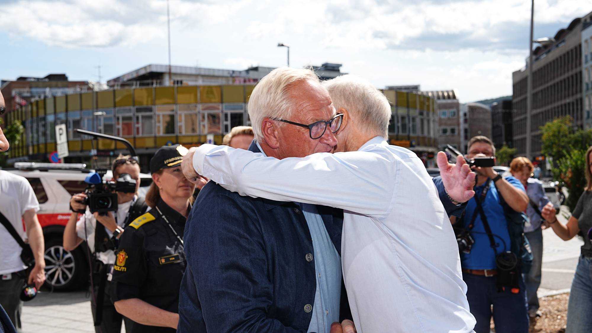 To menn omfavner hverandre utendørs i en gate, omgitt av pressefolk og en politibetjent. (Bildebeskrivelsen er laget av en KI-tjeneste)