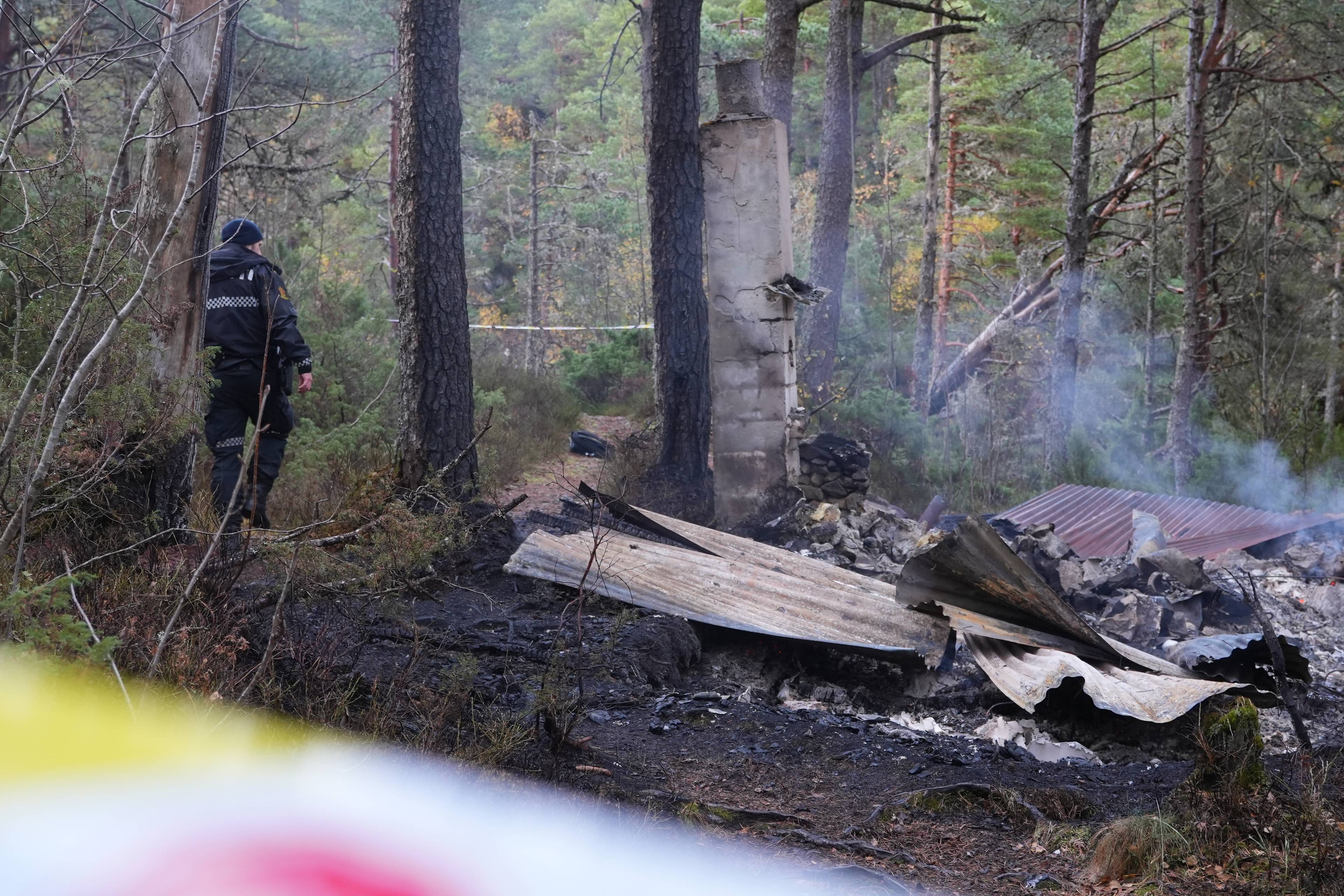 Politibetjent ved ruinene av en nedbrent bygning i skogen. Røyk stiger opp fra branntomten.