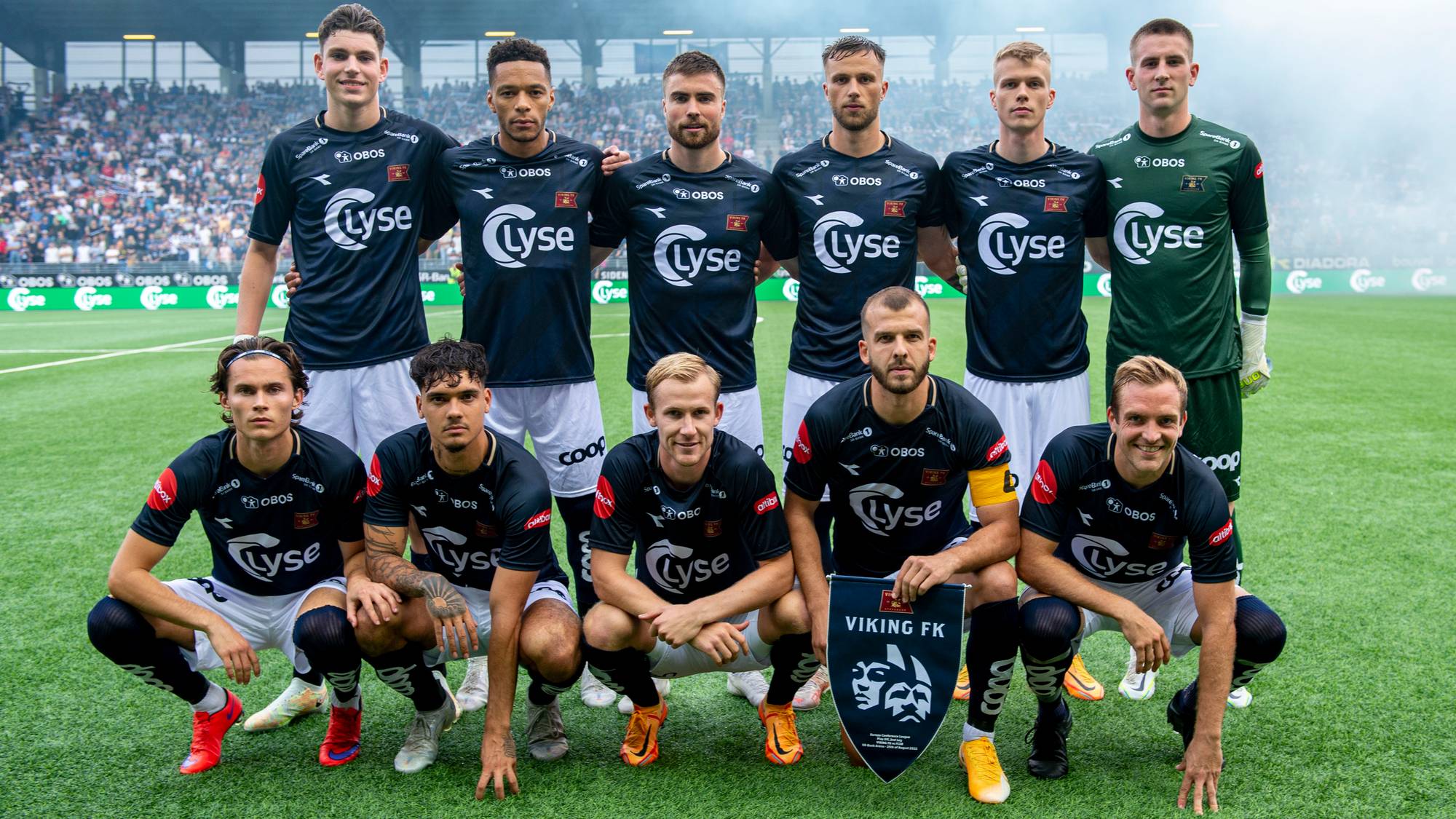 Elve fotballspillere poserer for lagbilde på en gressbane. Alle er ikledd mørke drakter med sponsorlogoer og hvite shorts. To spillere i forgrunnen holder et lite fanebanner med teksten "VIKING FK". I bakgrunnen er tribuner med tilskuere og et gjerde. (Bildebeskrivelsen er laget av en KI-tjeneste)