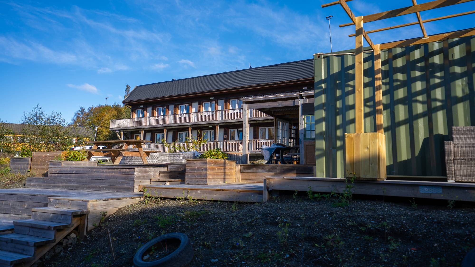 Bygning med terrasse og en grønn container under åpen himmel. (Bildebeskrivelsen er laget av en KI-tjeneste)