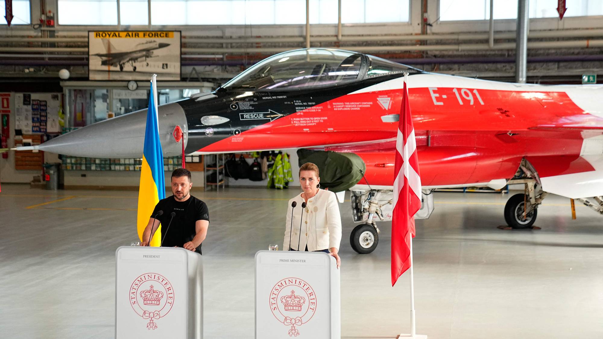 Mette Frederiksen og Zelenskyj framfor eit jagarfly. Dei står ved to podium med eit ukrainsk og eit dansk flagg. 