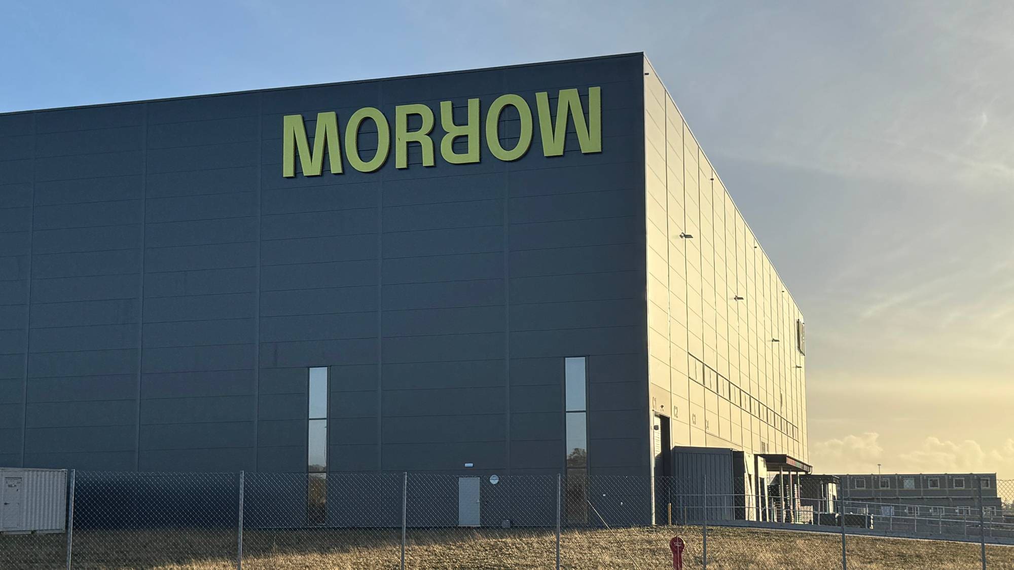 En stor, mørkegrå industribygning har et stort, gult skilt med teksten "MOROW". Bygningen har flere vinduer og en lasterampe. Foran bygningen er det et metallgjerde og tørt gress. (Bildebeskrivelsen er laget av en KI-tjeneste)