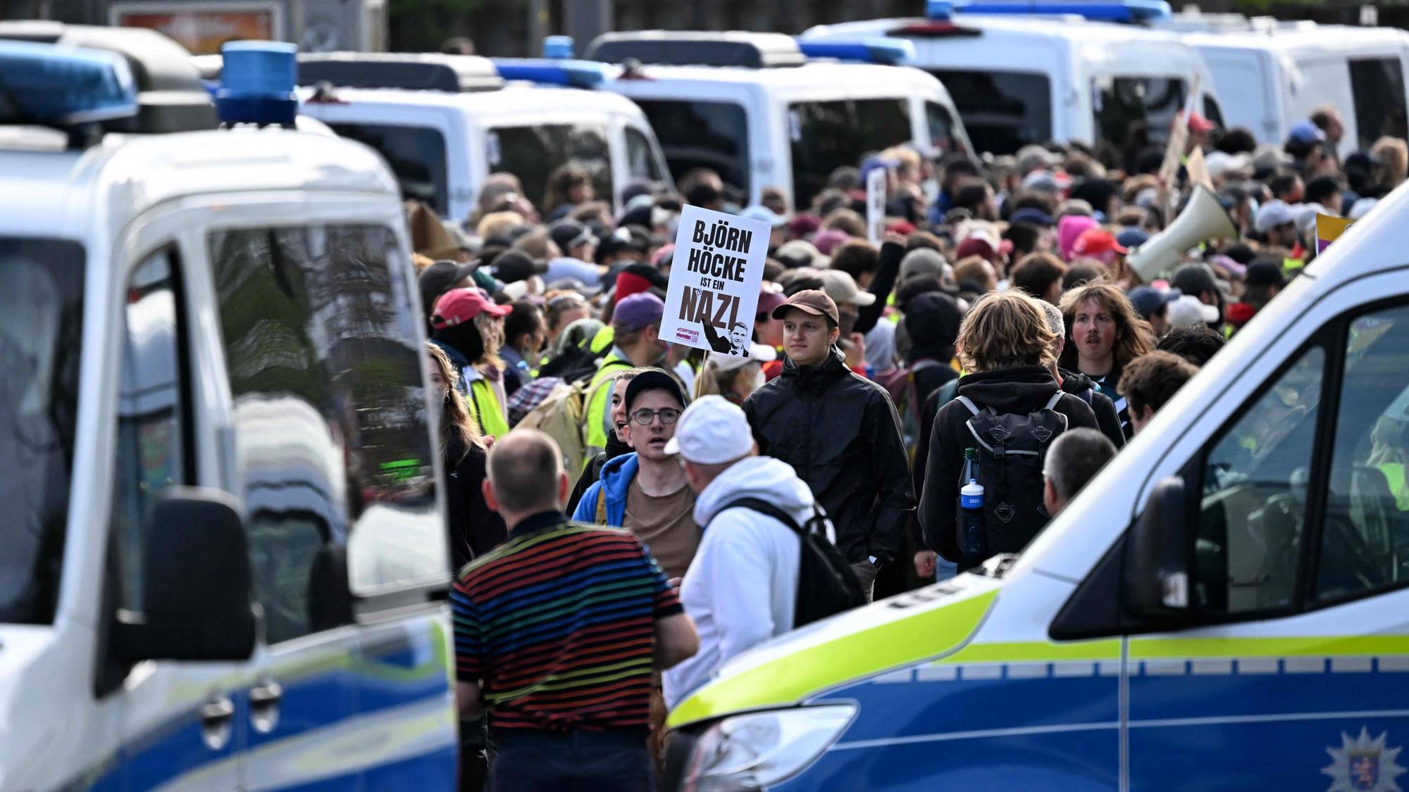 Demonstranter holder et skilt med tekst om Björn Höcke omgitt av politibiler. (Bildebeskrivelsen er laget av en KI-tjeneste)
