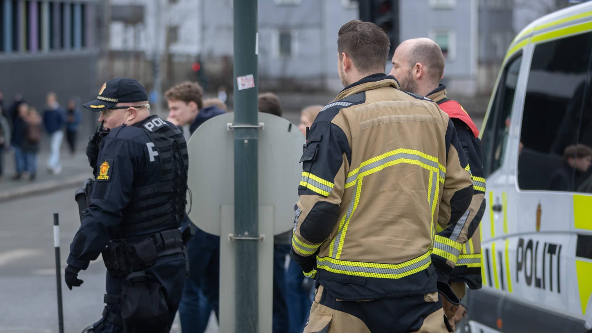 To politimenn og to brannkonstabler står ved en politibil. De kikker alle mot venstre. Politimannen snakker i kommunikasjonsutstyret. Vi ser mye folk i bakgrunnen