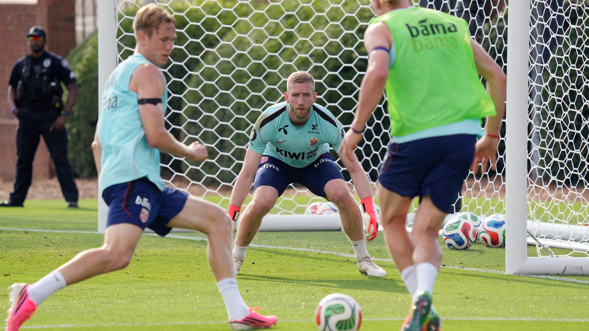 Tre fotballspillere trener foran et mål. En keeper står i målet og gjør seg klar til å ta imot ballen. To andre spillere er i bevegelse på banen. Bildet viser en fotballtrening. (Bildebeskrivelsen er laget av en KI-tjeneste)