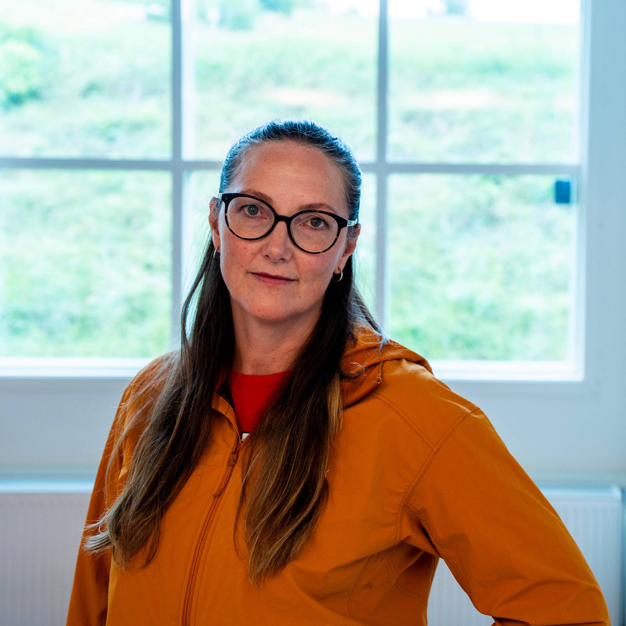 Kvinne med briller og oransje jakke står foran et vindu innendørs. (Bildebeskrivelsen er laget av en KI-tjeneste)