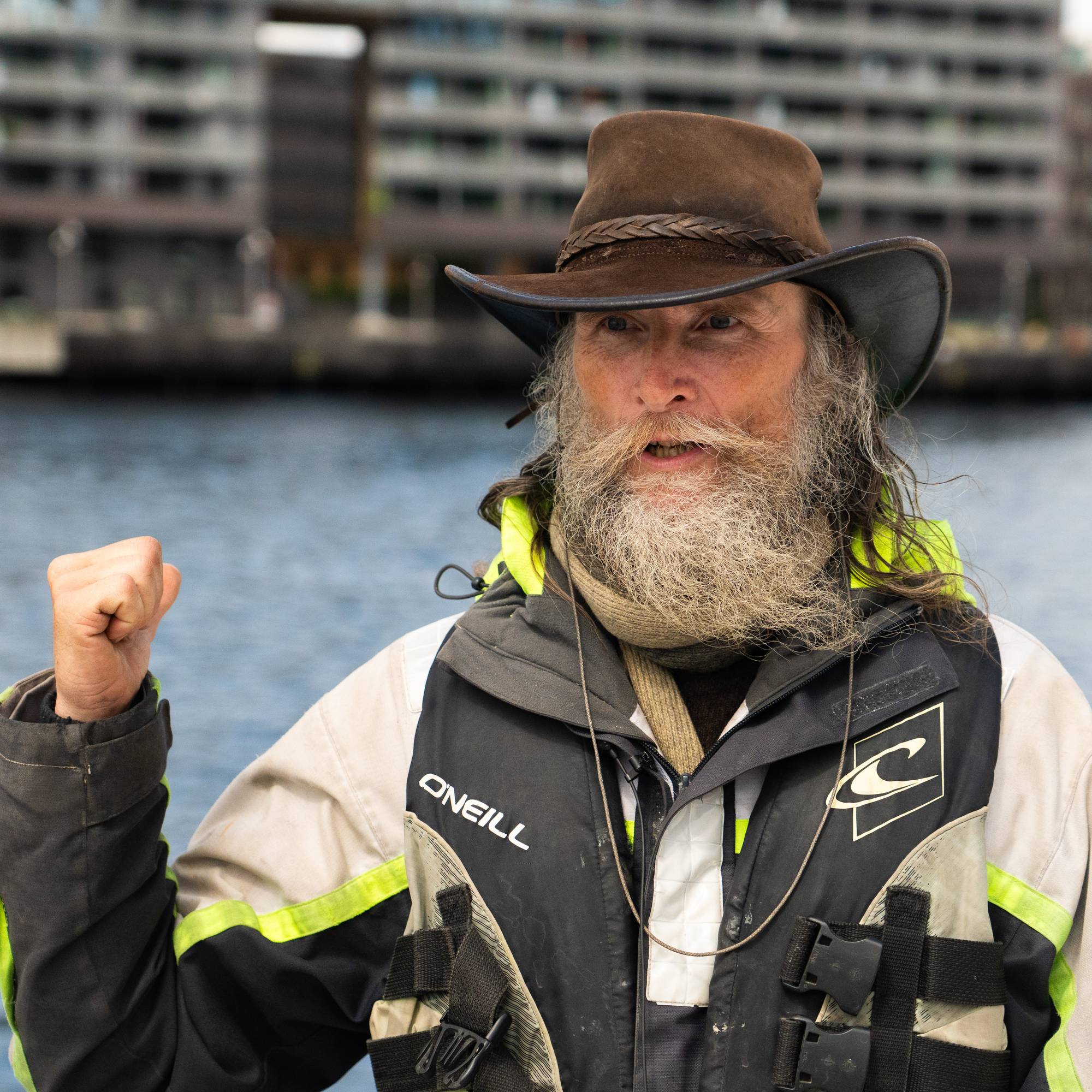 Mann med langt skjegg, cowboyhatt, seilerjakke og flytevest med sjøen og Sørenga i bakgrunnen
