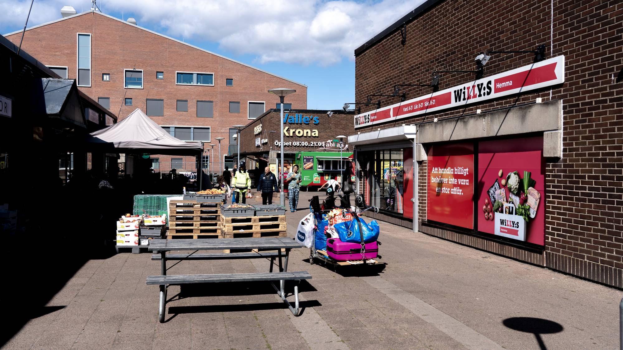 Personer går over en plass med butikker og markedsboder i dagslys. (Bildebeskrivelsen er laget av en KI-tjeneste)