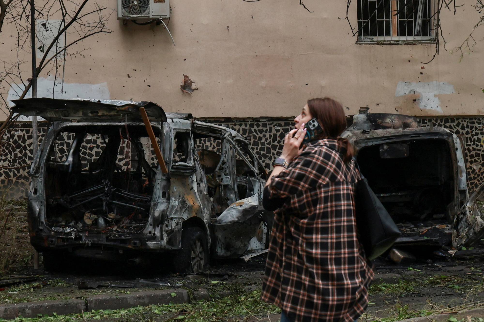 En kvinne snakker på telefon foran to utbrente biler. Bygningen bak dem er skadet, med tydelige merker etter eksplosjoner på veggen. En av bilene har bagasjerommet åpent. Omgivelsene ser kaotiske ut. (Bildebeskrivelsen er laget av en KI-tjeneste)