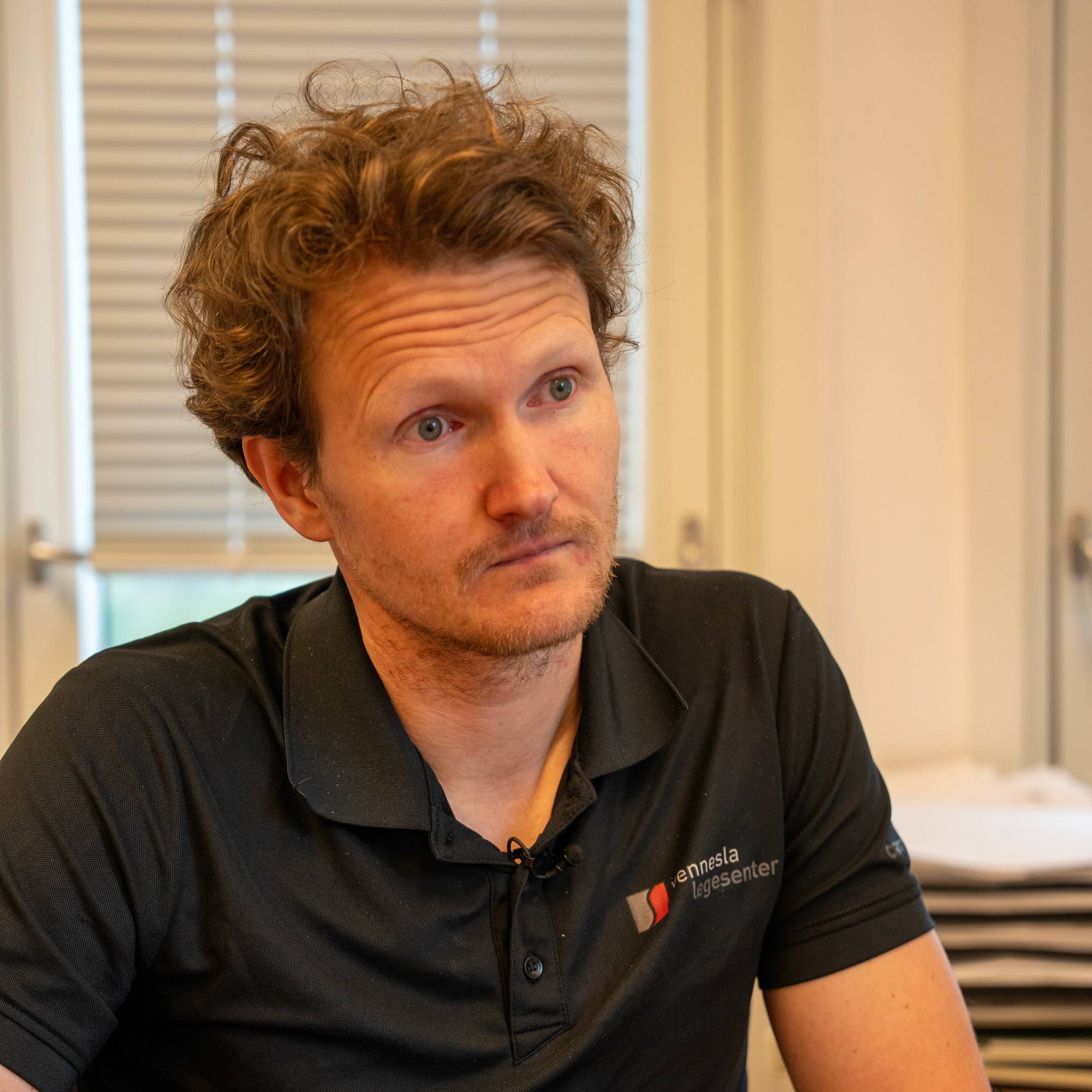En mann med krøllete brunt hår sitter og snakker. Han har på seg en sort polo-skjorte med logo fra "Sennsnes legesenter". Mannen har hendene foldet foran seg. Bakgrunnen viser et vindu med persienner og et skrivebord. (Bildebeskrivelsen er laget av en KI-tjeneste)