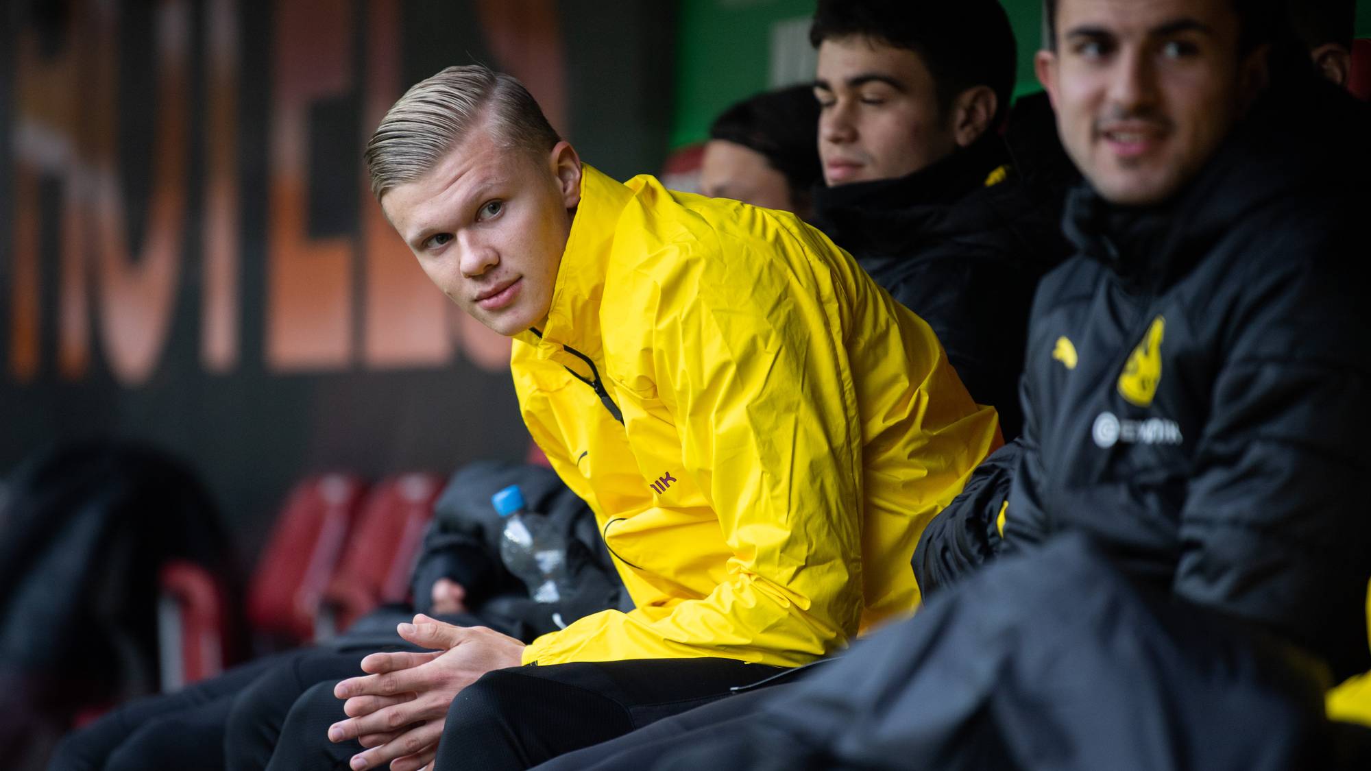 Erling Braut Haaland sitter på spillerbenken i gul treningsjakke sammen med lagkamerater. (Bildebeskrivelsen er laget av en KI-tjeneste)