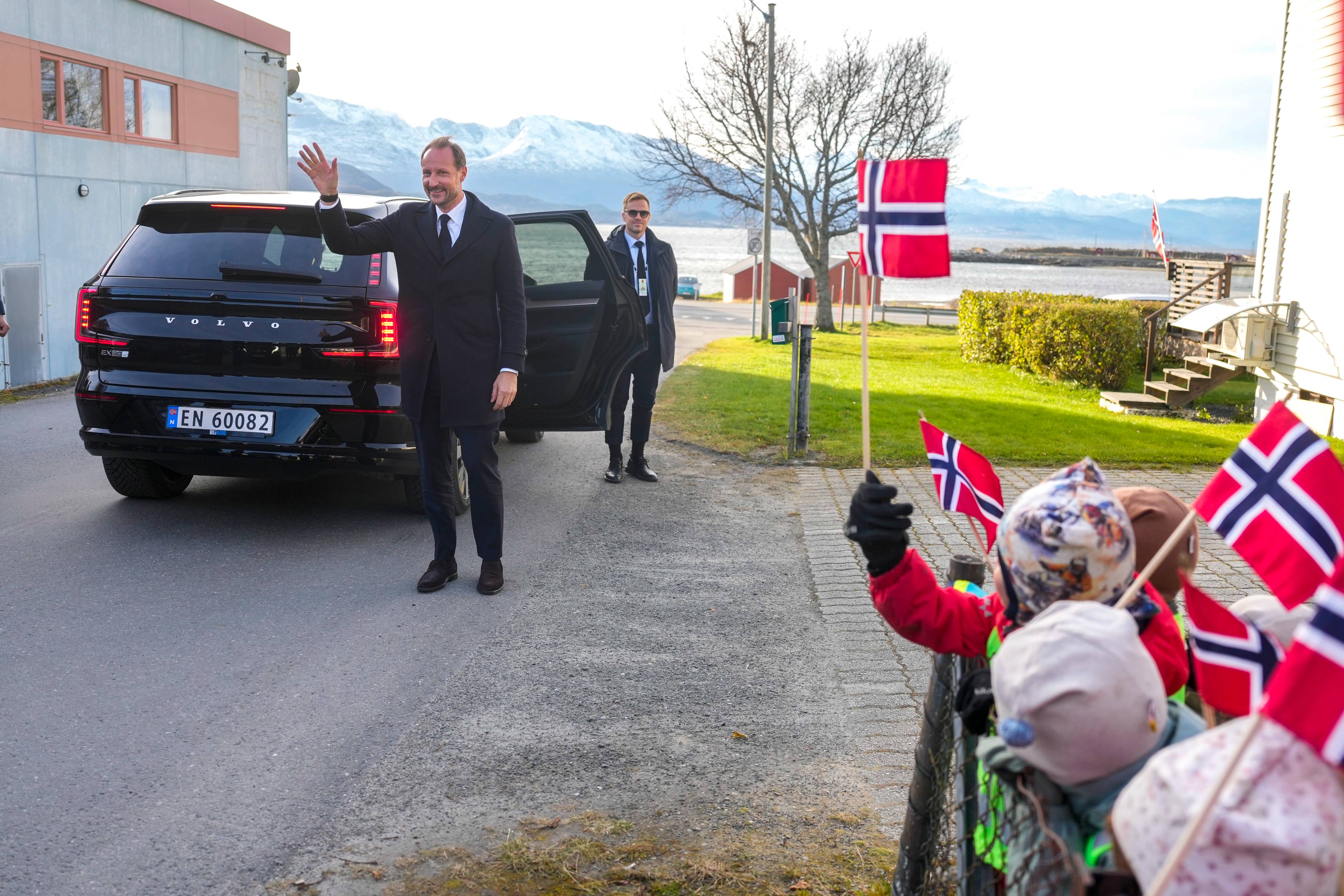 Kronprins Haakon vinker til barn som holder norske flagg ved en bil. (Bildebeskrivelsen er laget av en KI-tjeneste)