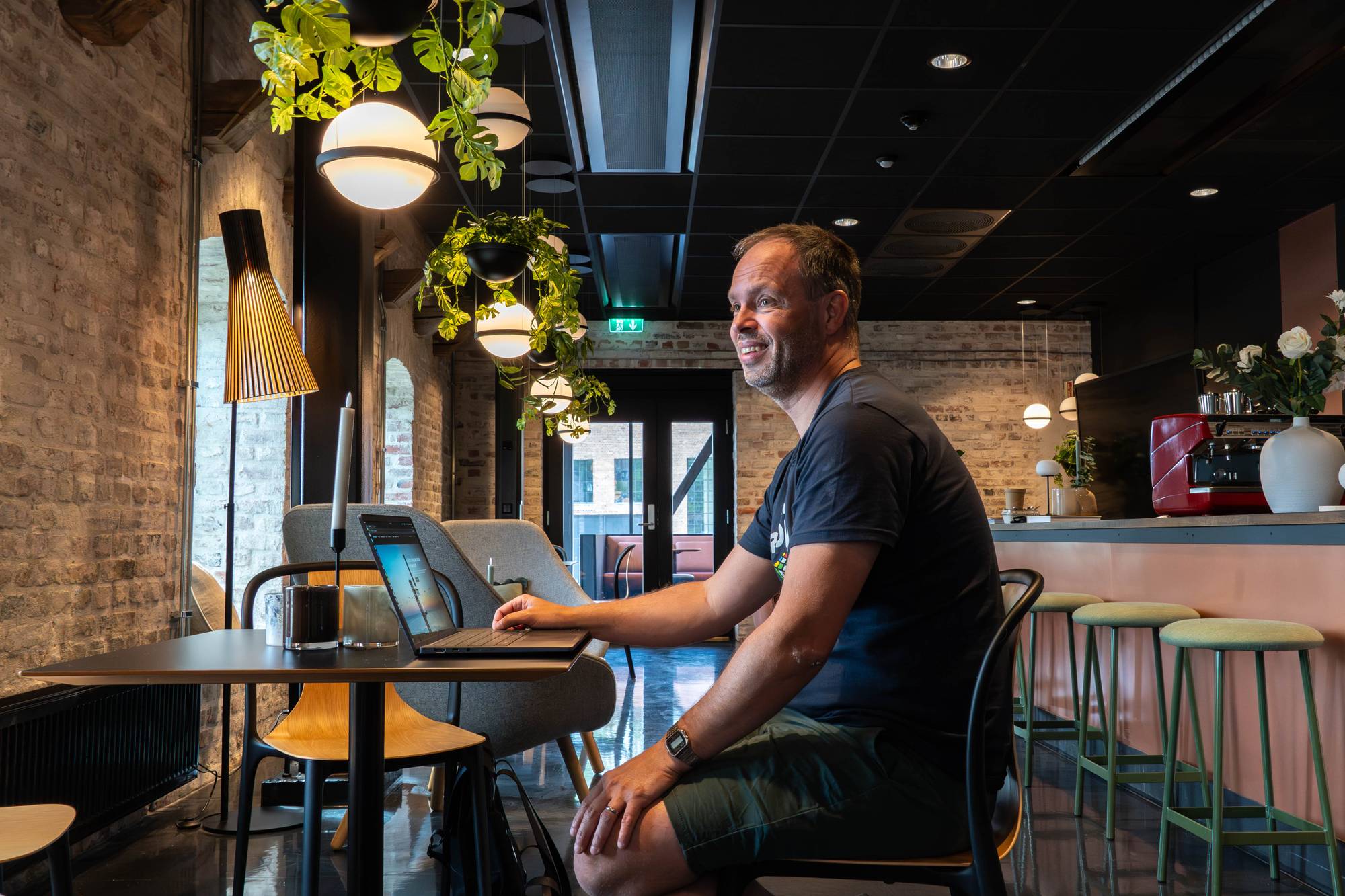 En mann sitter ved et bord med en bærbar PC foran seg. Han ser mot høyre og smiler. Bak ham henger planter fra taket. Rommet har en kafé- eller loungeaktig atmosfære med mursteinsvegger og moderne belysning. (Bildebeskrivelsen er laget av en KI-tjeneste)