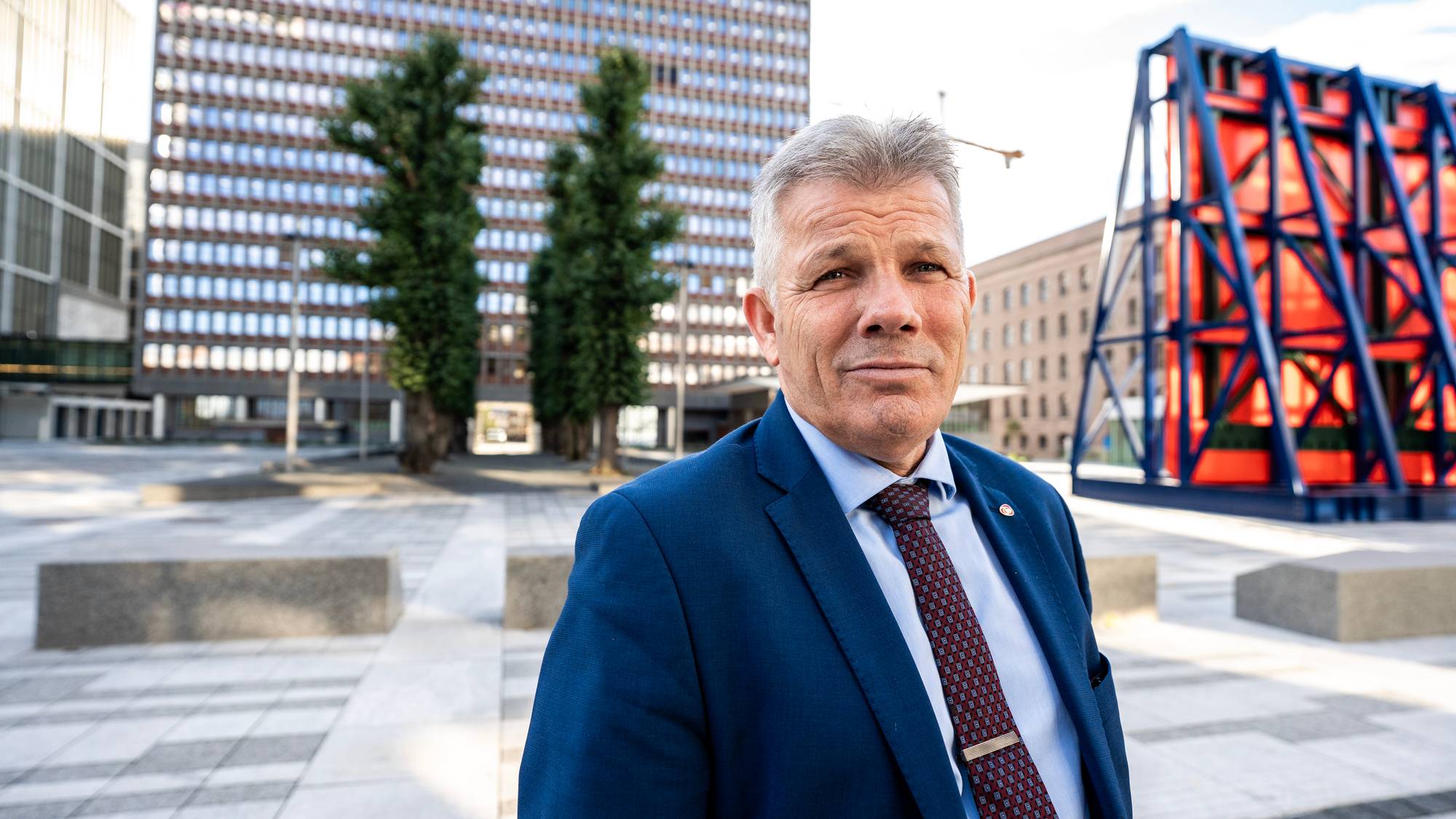Mann i dress og slips står utendørs foran Høyblokka i Regjeringskvartalet i Oslo. (Bildebeskrivelsen er laget av en KI-tjeneste)