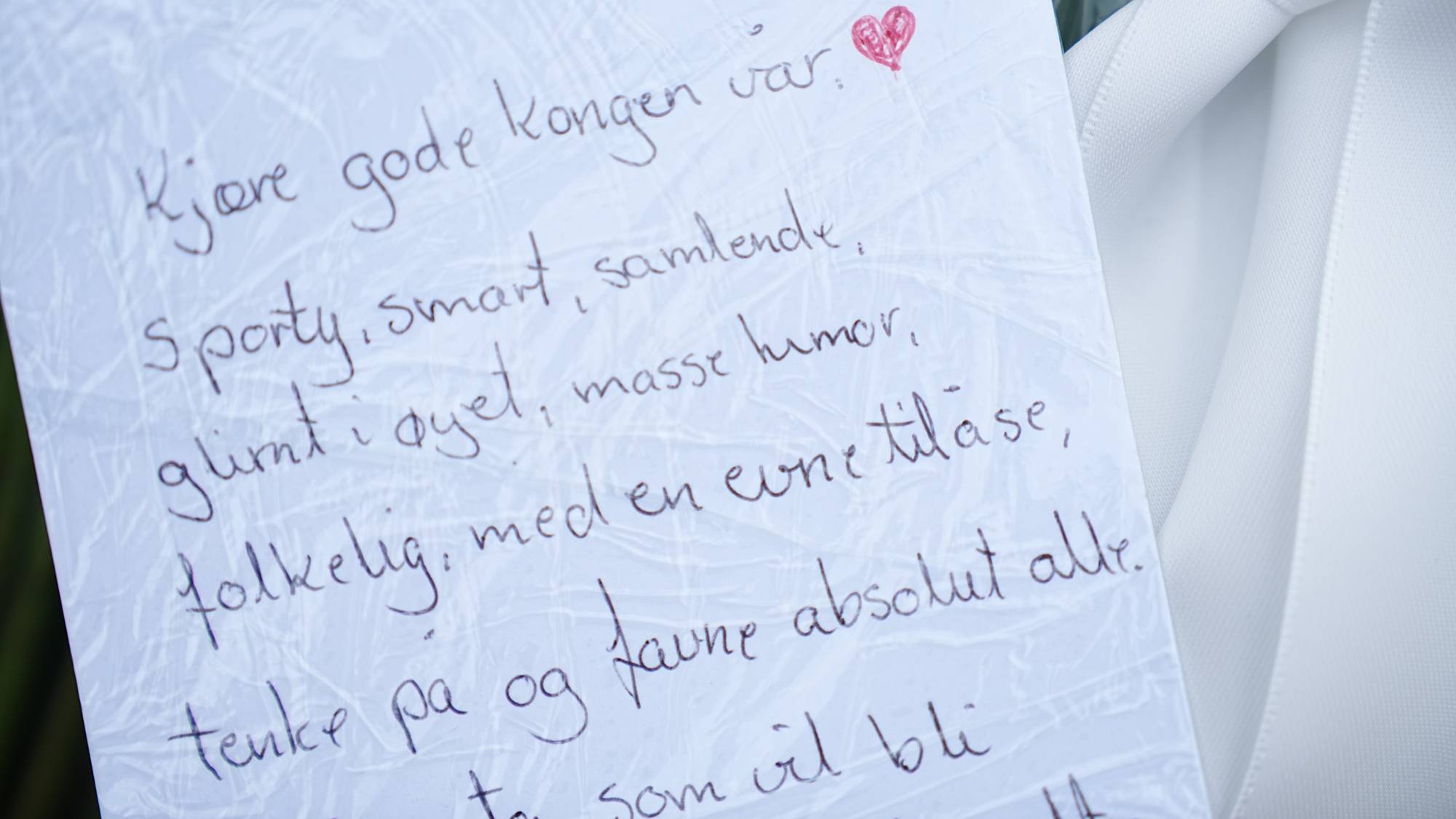 Håndskrevet hilsen til kongen festet til en blomsterbukett med hvitt bånd. (Bildebeskrivelsen er laget av en KI-tjeneste)