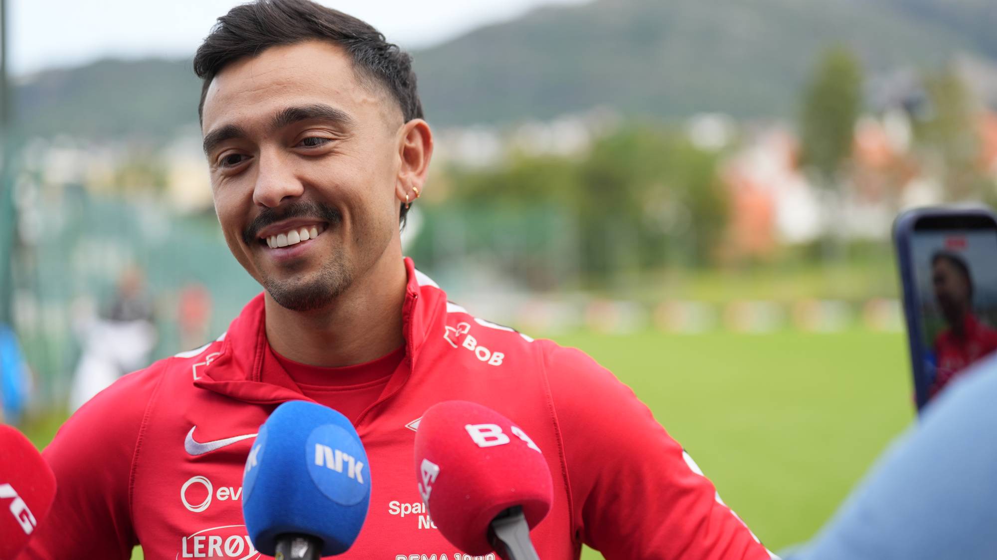 Fotballspiller smiler mens han blir intervjuet av journalister med mikrofoner. 