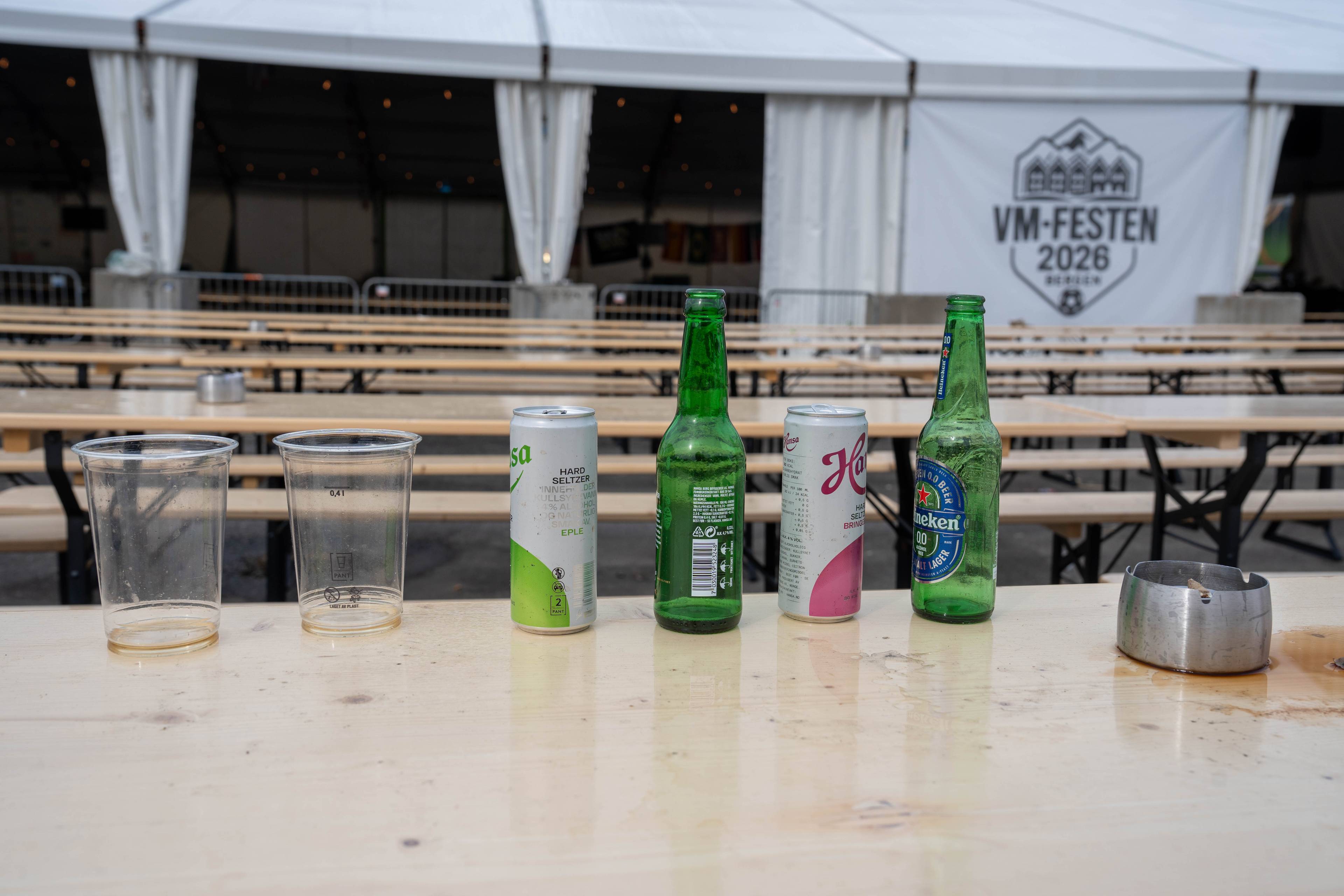 To tomme plastbeger står på et bord føre ein rad med tomme bord og stoler. Mellom begrene står to boksar og to flasker, ein av dei med etiketten «Heineken». Bak bordet er et stort kvitt telt. (Bildebeskrivelsen er laget av ein KI-teneste)