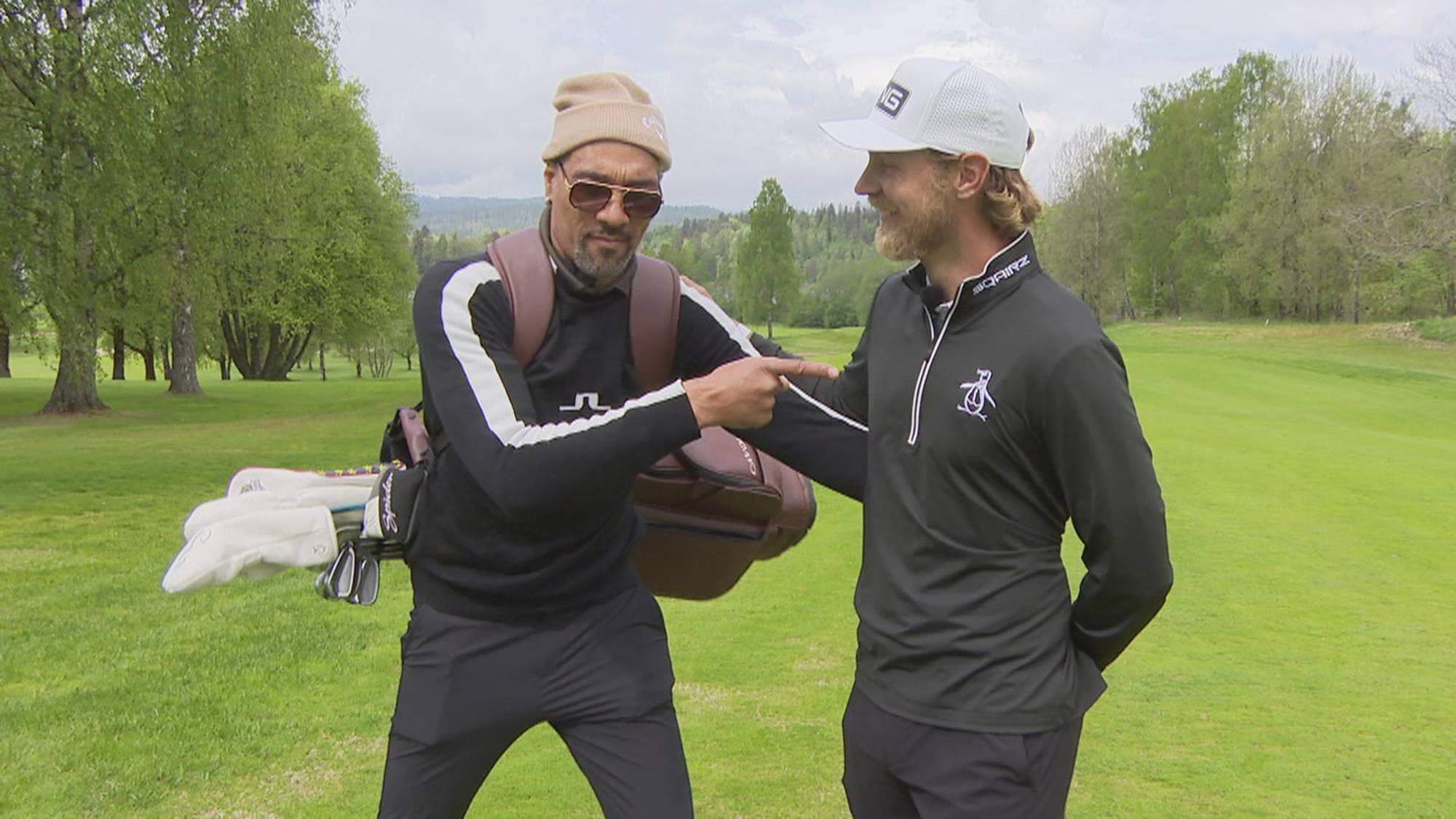 to menn på en golfbane. Han ene har på lue og peker på han andre som har på caps. De er begge glade 