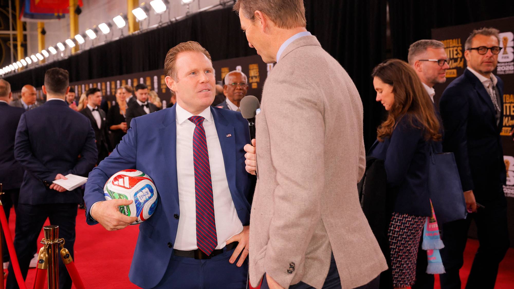 En mann i blå dress med et fotball er intervjuet på et rødt teppe. En annen mann i en lys brun dress holder en mikrofon. Bak dem er det flere gjester og et banner for FIFA World Cup. (Bildebeskrivelsen er laget av en KI-tjeneste)