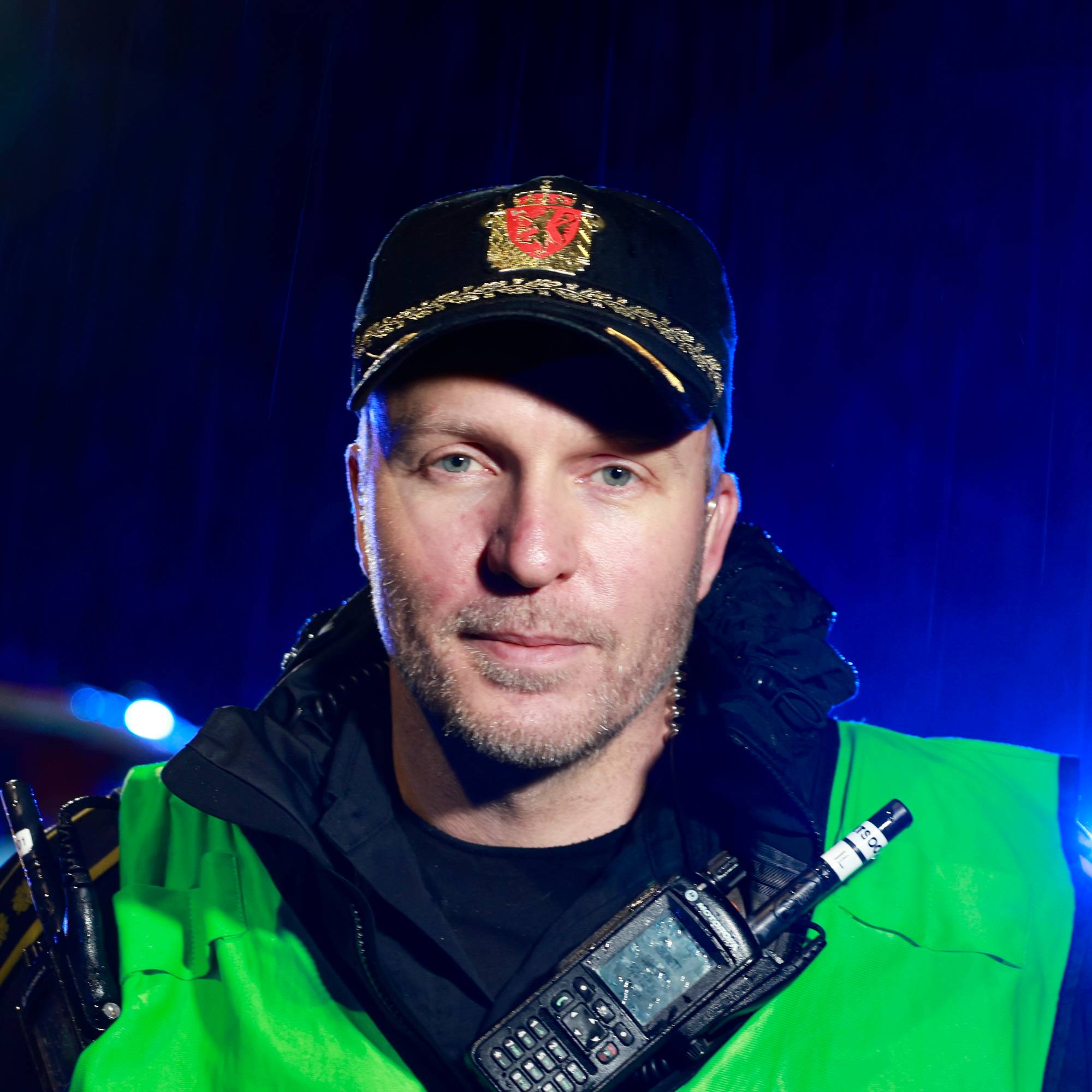 Politimann med caps, grønn refleksvest og hendene på hoftene. Mørkt og blått lys skimtes i bakgrunnen. 
