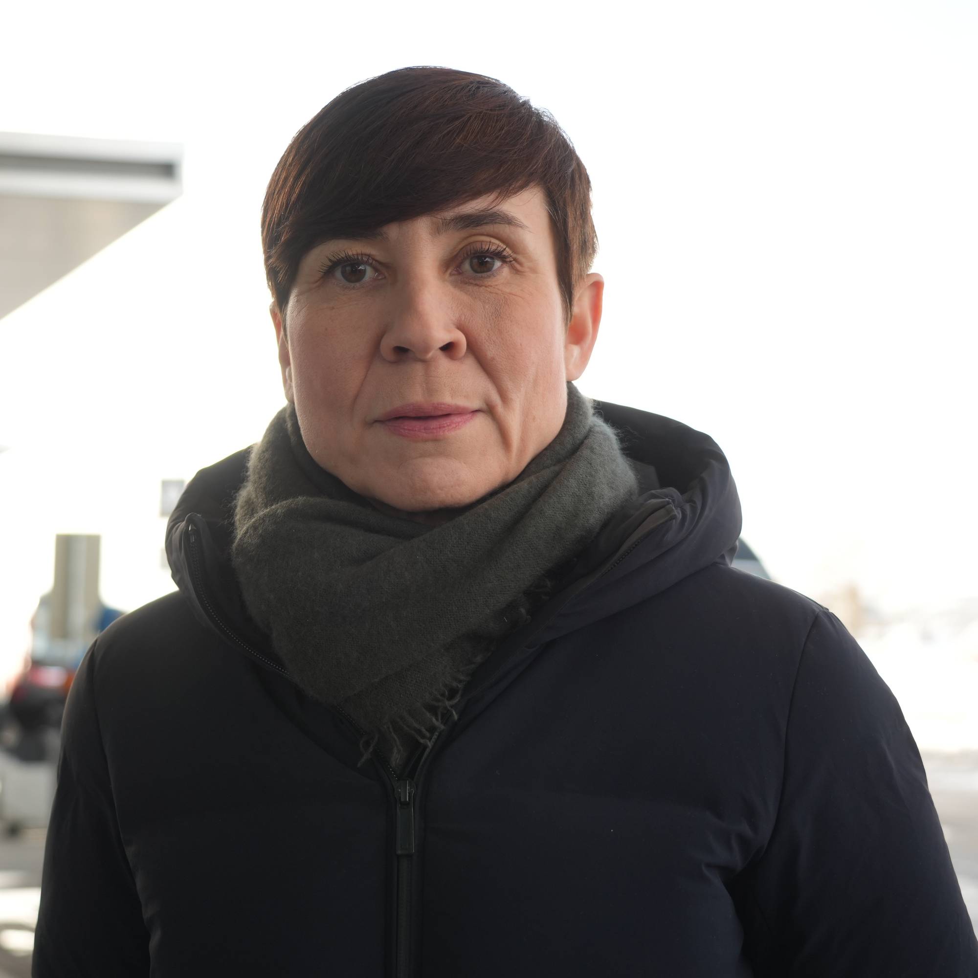 En kvinne med mørkt, kort hår står ute foran en bygning. Hun har på seg en mørk boblejakke og et mørkegrått skjerf. Bak henne er en moderne bygningsfasade med store glassflater. I bakgrunnen ser man uskarpe biler og en gul kiosk. Lyset er sterkt og himmelen er lys. (Bildebeskrivelsen er laget av en KI-tjeneste)