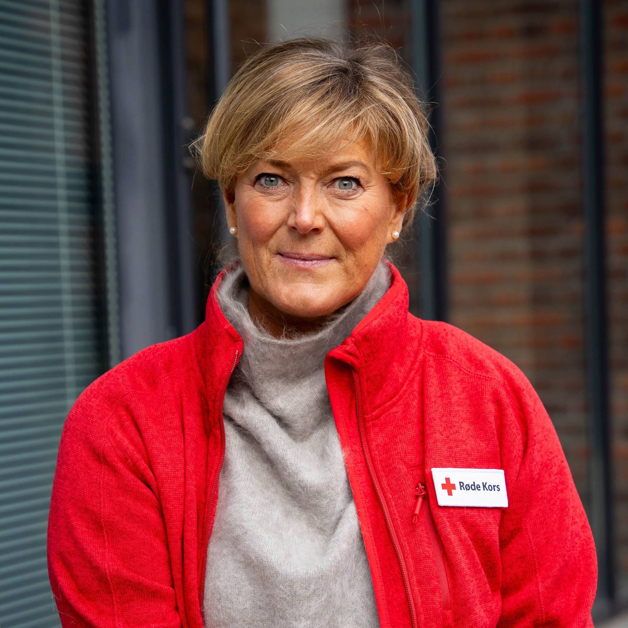 Kvinne med blondt hår i rød Røde Kors-jakke ser inn i kameraet. (Bildebeskrivelsen er laget av en KI-tjeneste)