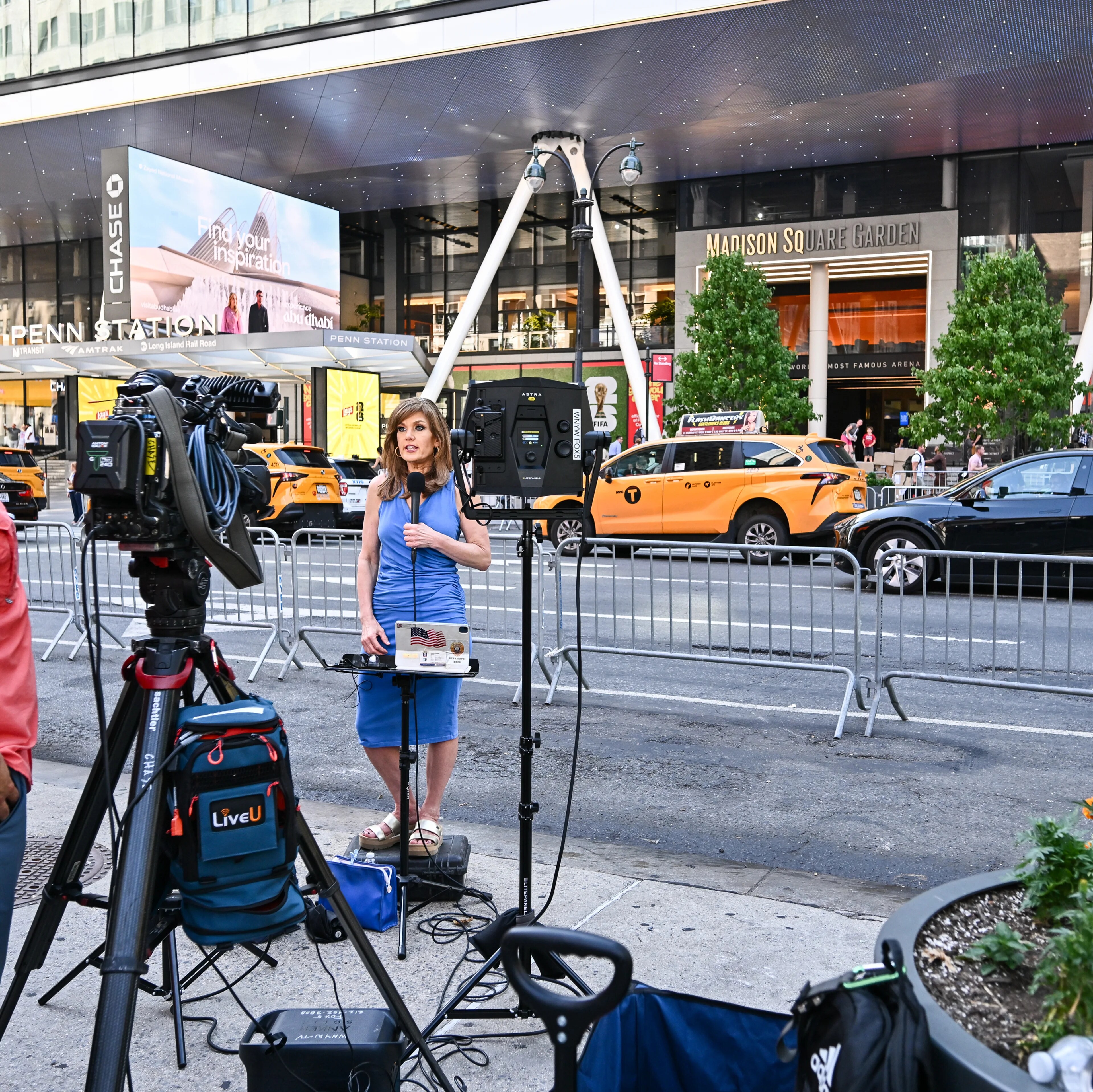 Kvinne i blå kjole rapporterer fra Madison Square Garden mens en fotograf filmer. (Bildebeskrivelsen er laget av en KI-tjeneste)