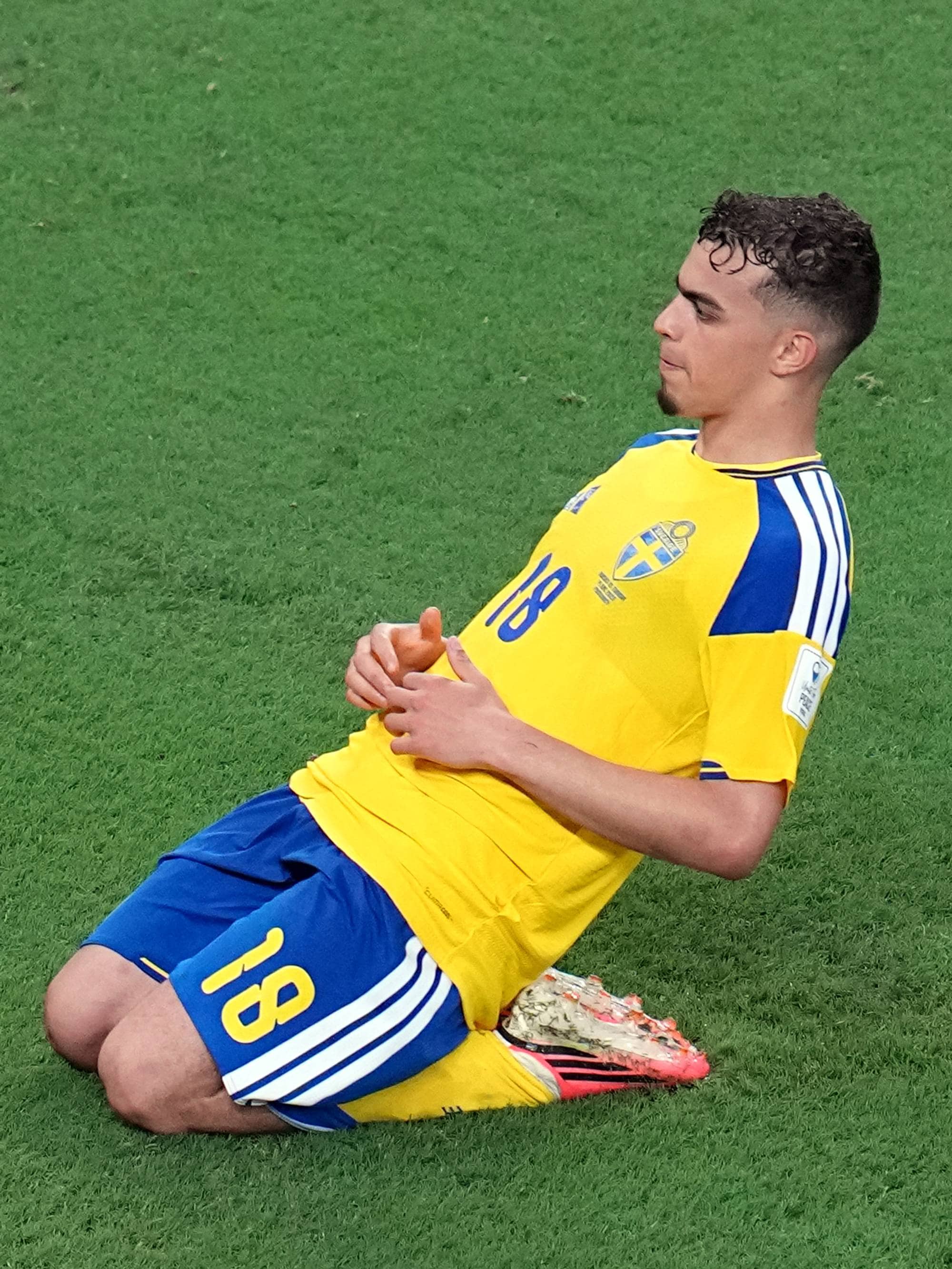 Fotballspiller i gul drakt med nummer 18 på kne på gressbane.
Spilleren har blå shorts med hvite striper.
Han ser mot høyre med hendene på brystet.
Fotballbanen er grønn med hvite markeringer. (Bildebeskrivelsen er laget av en KI-tjeneste)