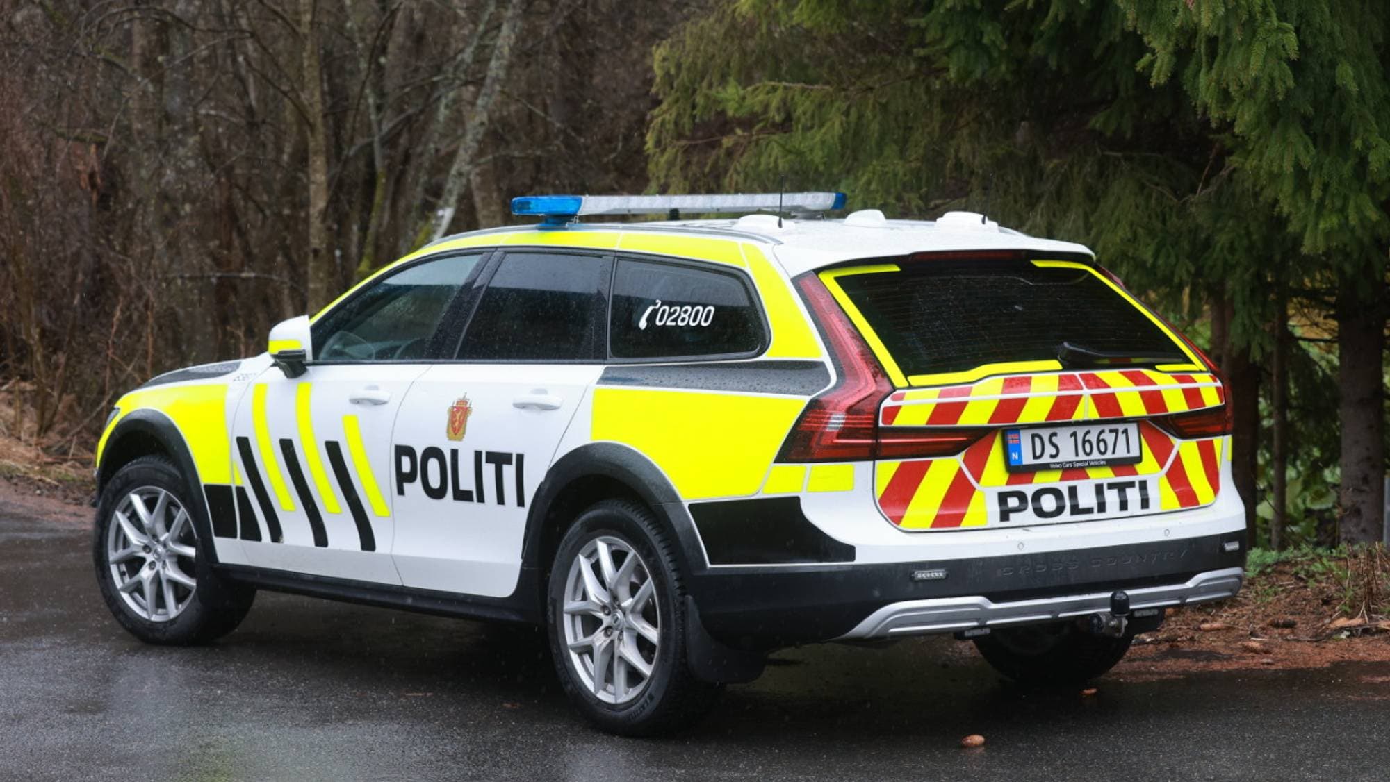 En politibil står parkert på en våt vei. Bilen er hvit med gule og svarte markeringer, og teksten "POLITI" er tydelig synlig. Blålys er aktivert på taket. I bakgrunnen er det trær og vegetasjon. (Bildebeskrivelsen er laget av en KI-tjeneste)