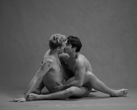 To unge menn sitter nakne ansikt til ansikt. Han ene har kort mørkt hår og han andre har kort blondt hår. De holder rundt hverandre og kysser.