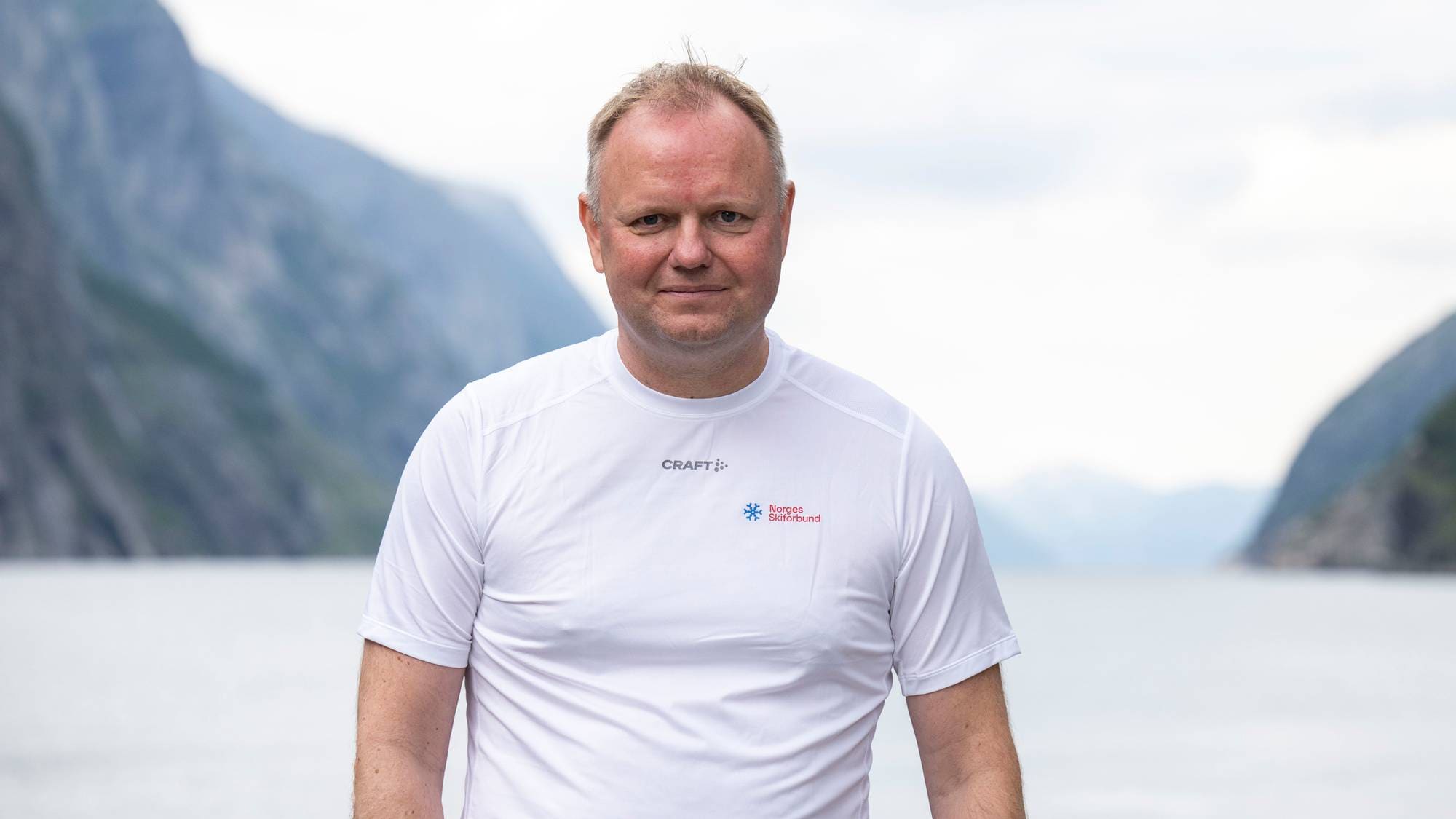 En mann i hvit t-skjorte står foran en fjord med bratte fjellsider i bakgrunnen. (Bildebeskrivelsen er laget av en KI-tjeneste)