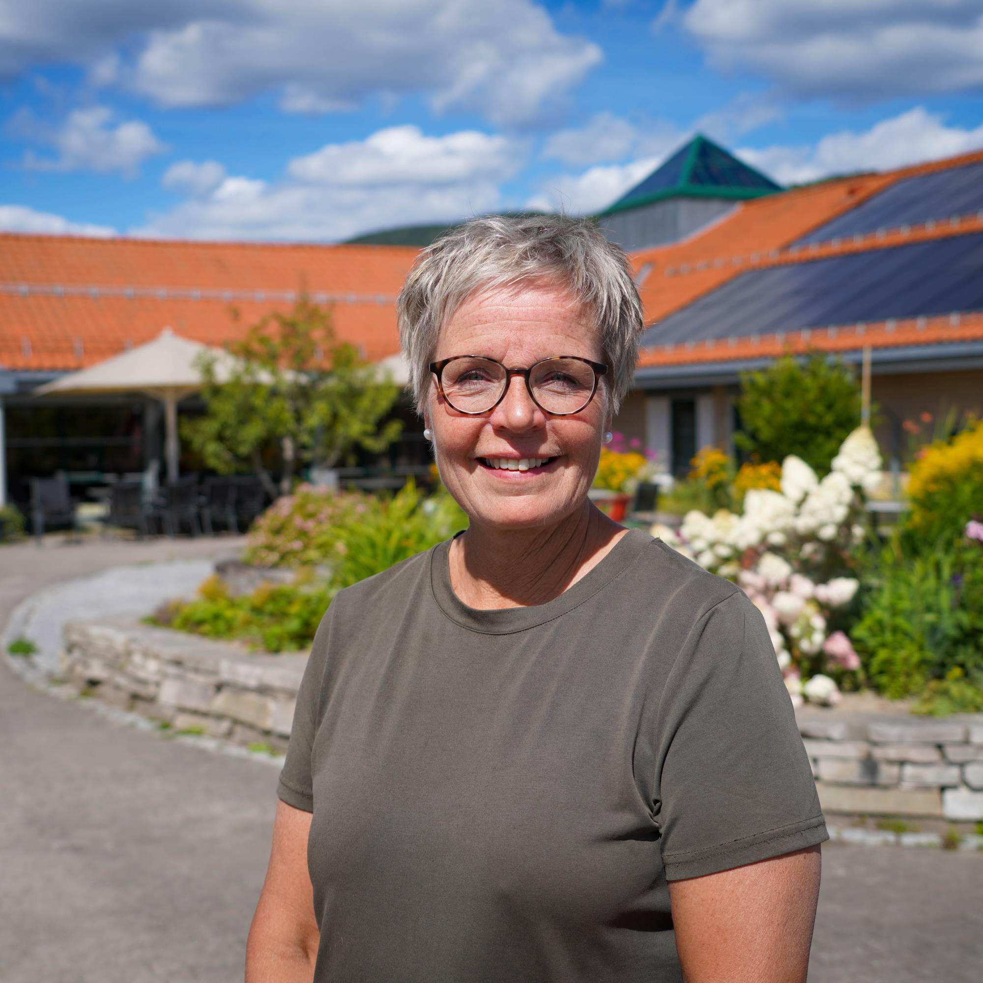 Kvinne med kort hår og briller smiler utendørs foran en bygning med hage. 