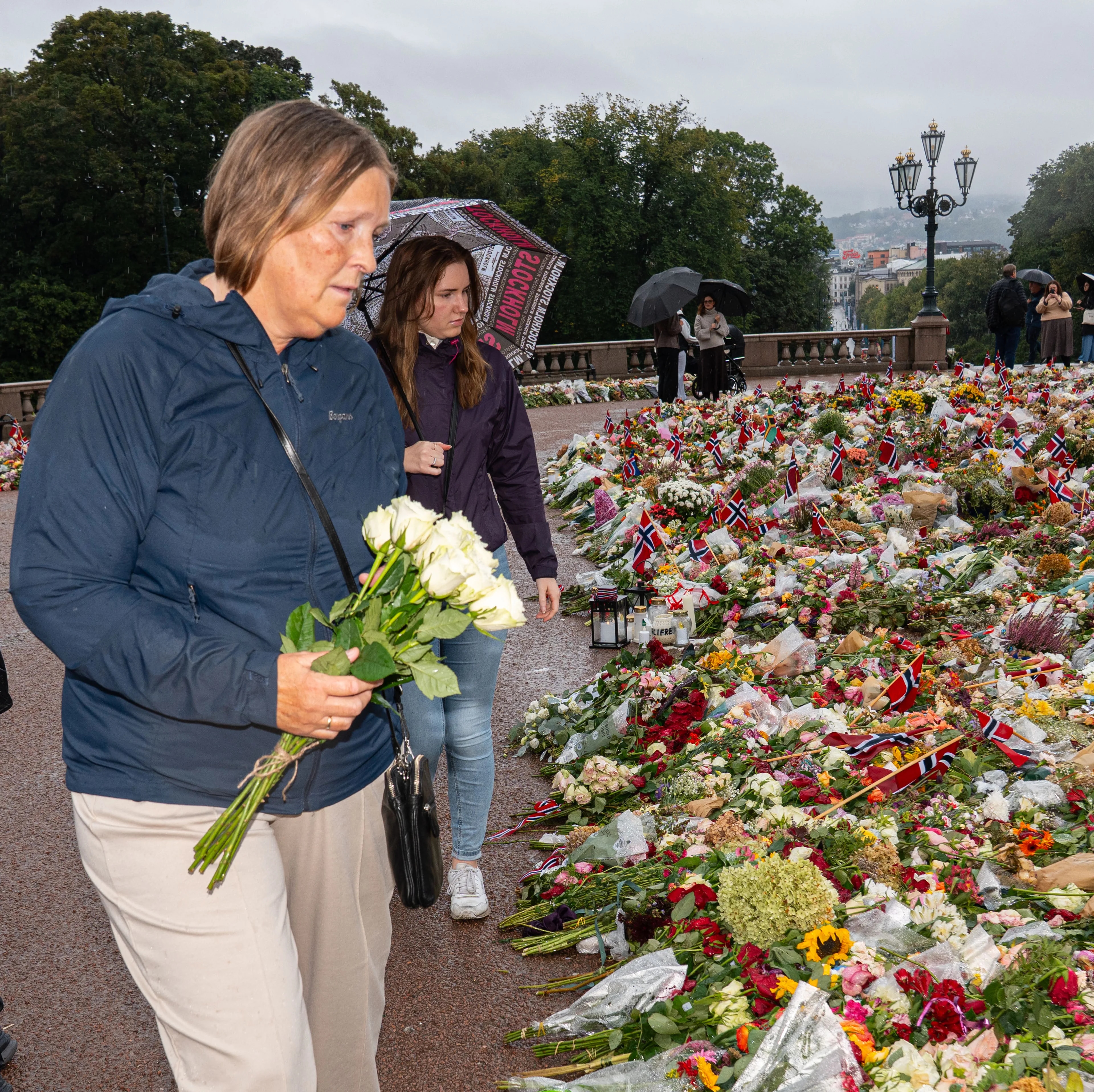 Kvinner legger ned blomster ved et stort minnested med blomsterhav og norske flagg i Oslo.