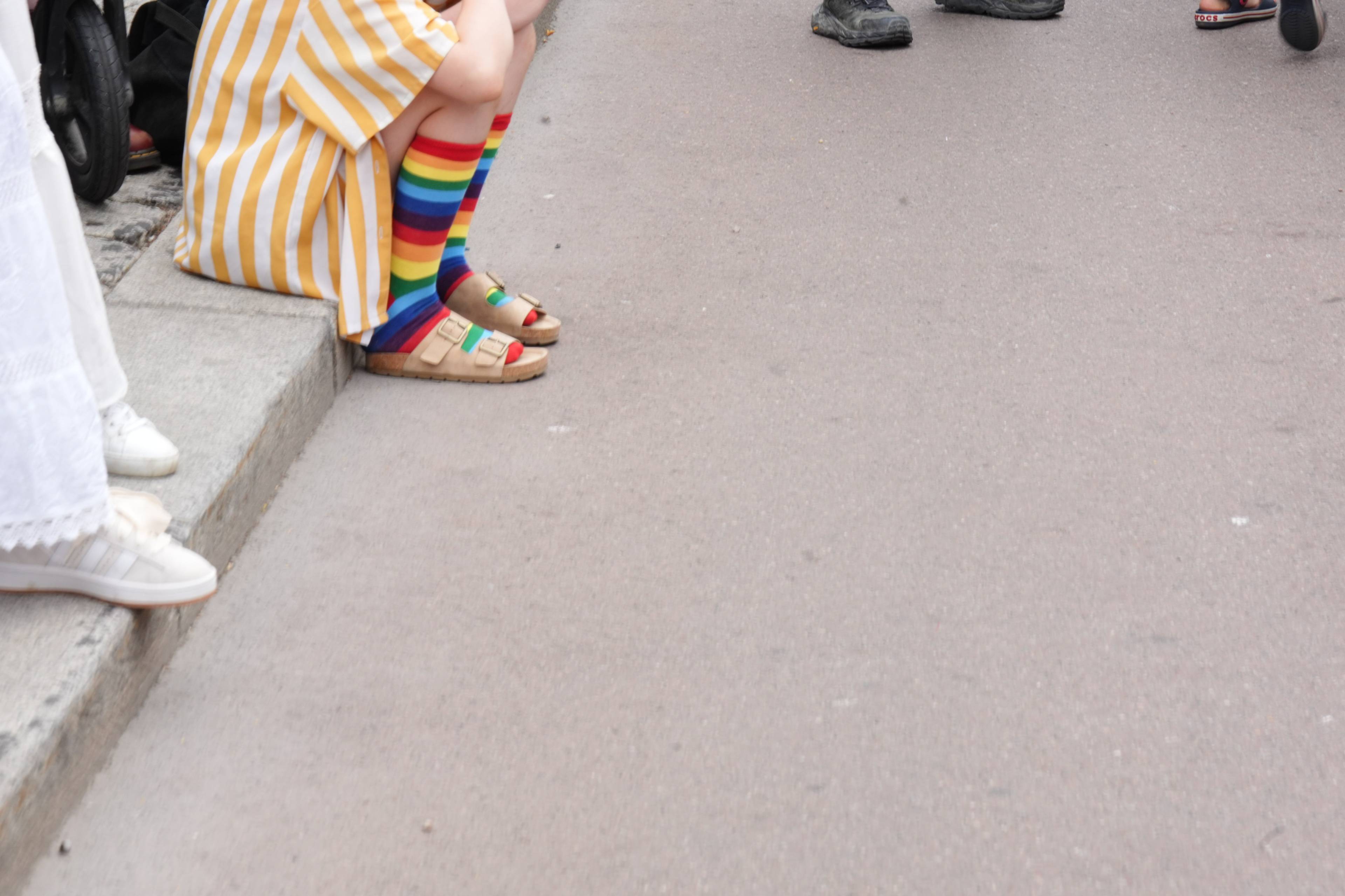 Person sitter på fortauskant med regnbuestripete sokker og sandaler. Andre personer står ved siden. (Bildebeskrivelsen er laget av en KI-tjeneste)