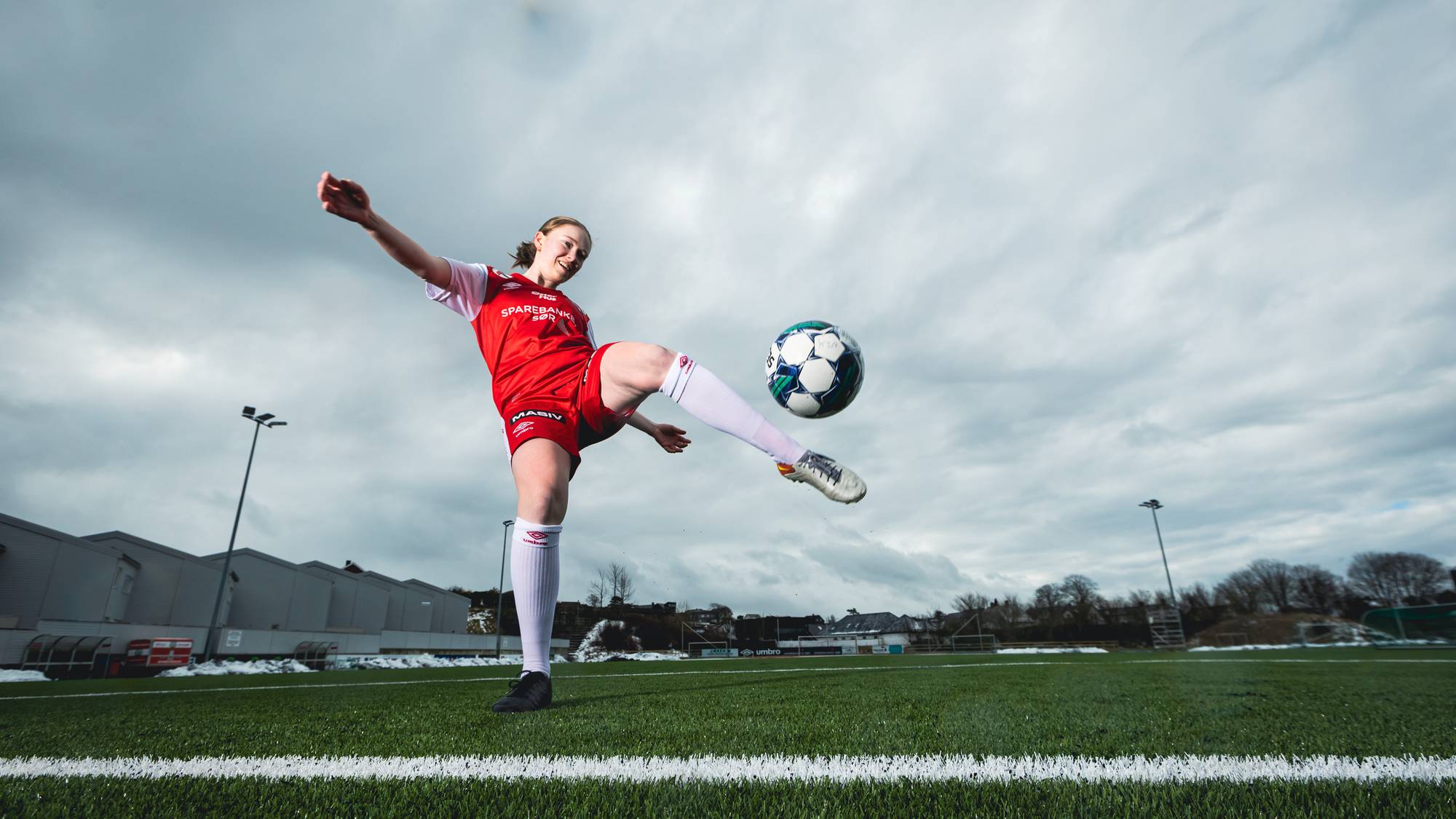 Bilde av Karina som sparker en fotball. Hun har på seg en rød og hvit fotballdrakt og er plassert på en fotballstadion. 