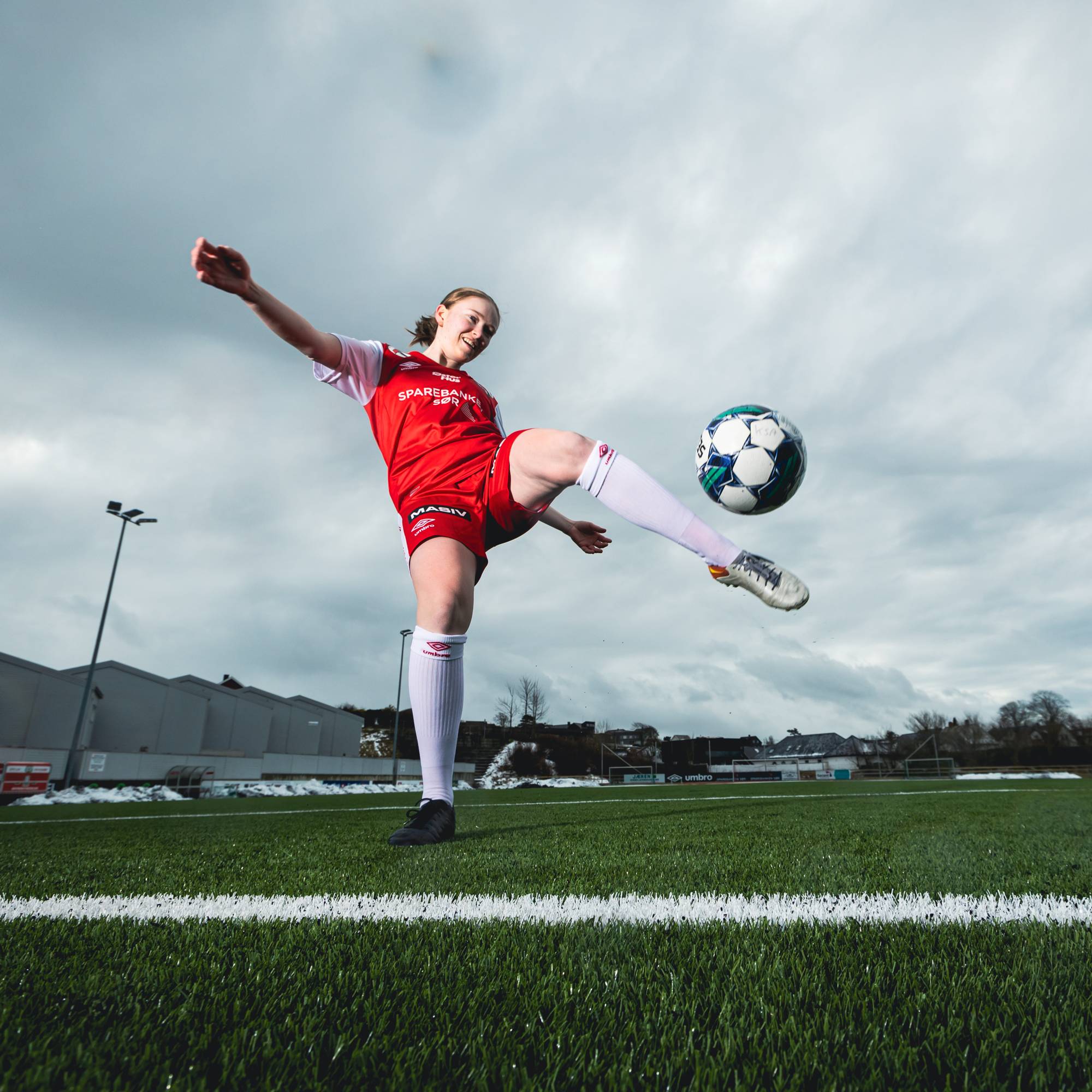 Bilde av Karina som sparker en fotball. Hun har på seg en rød og hvit fotballdrakt og er plassert på en fotballstadion. 
