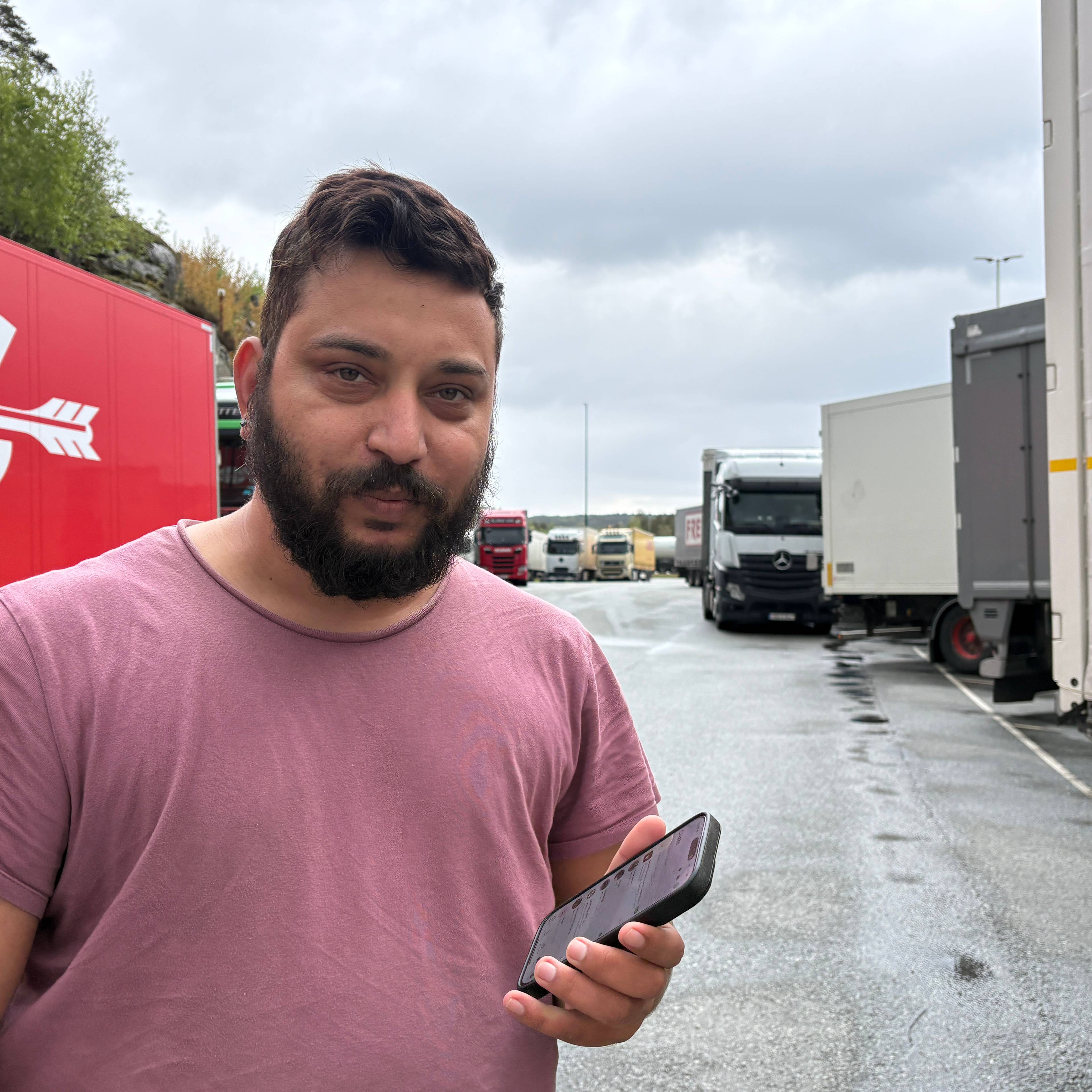 EIN mann med mørkt hår og skjegg, rødrosa T-skjorte står med mobil i handa, Han står mellom vogntog som er parkert på ein stor plass