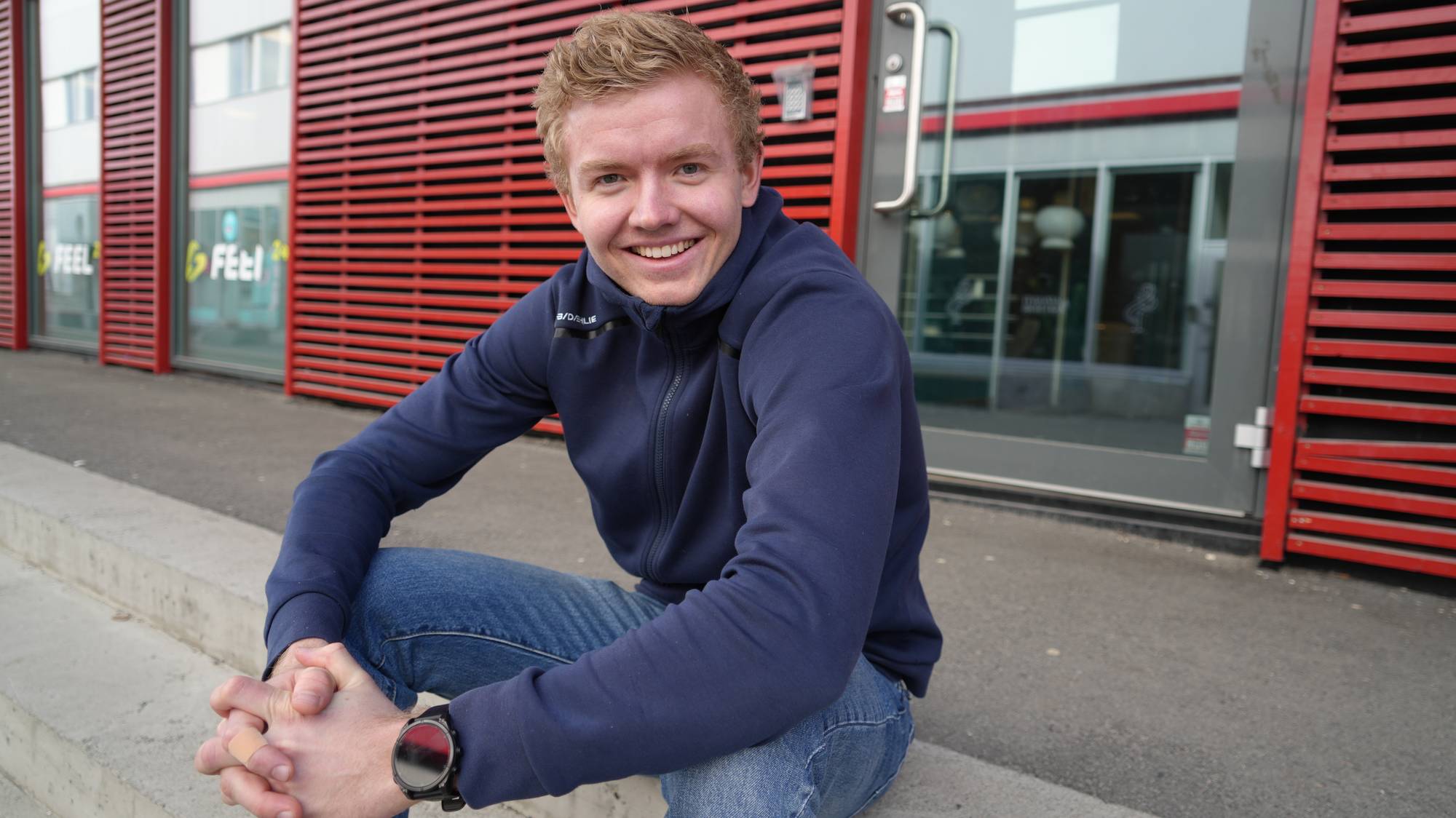 En ung smilende mann med lyst kort hår sittende på en betongtrapp