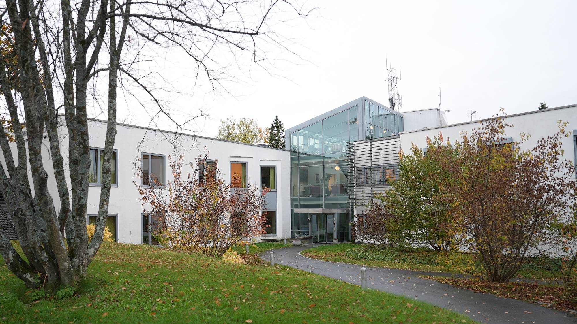 Moderne hvit bygning med glassfasade omgitt av en park med trær og en asfaltert sti. (Bildebeskrivelsen er laget av en KI-tjeneste)