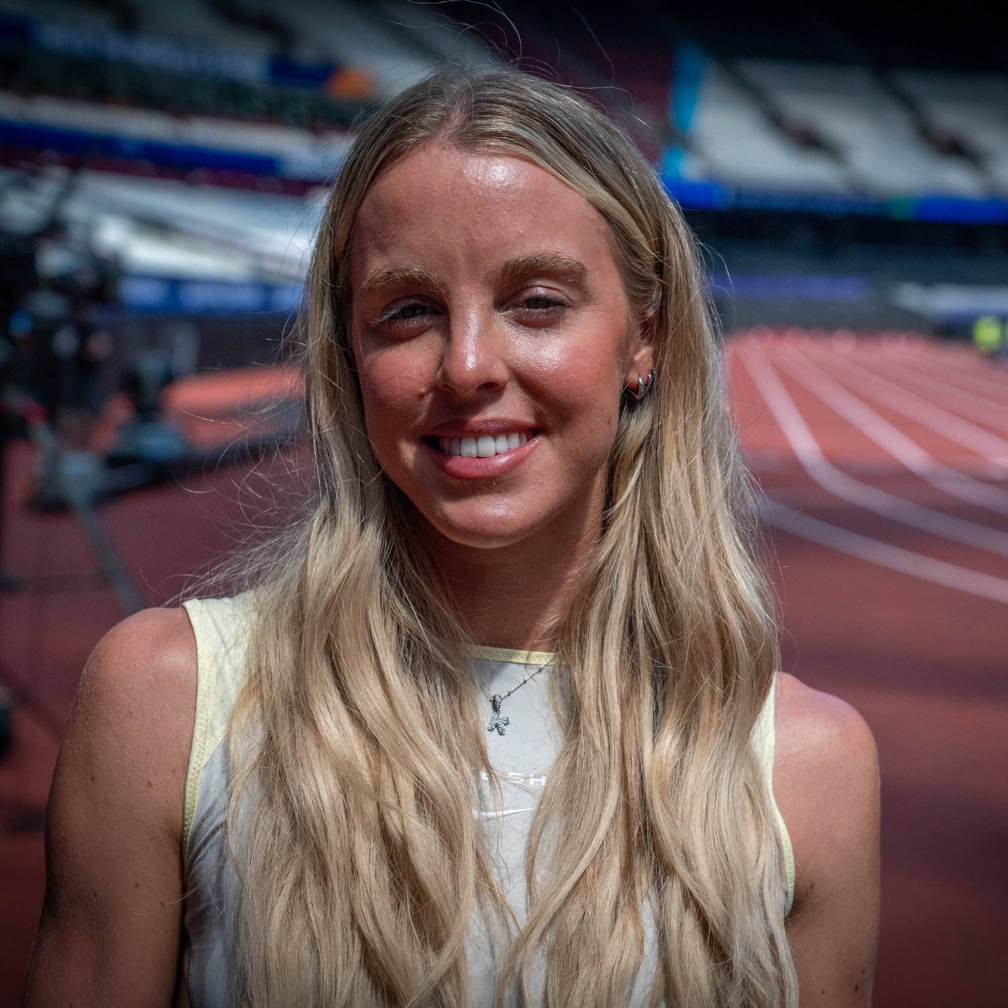Kvinne med blondt hår smiler foran en løpebane på en idrettsarena. (Bildebeskrivelsen er laget av en KI-tjeneste)