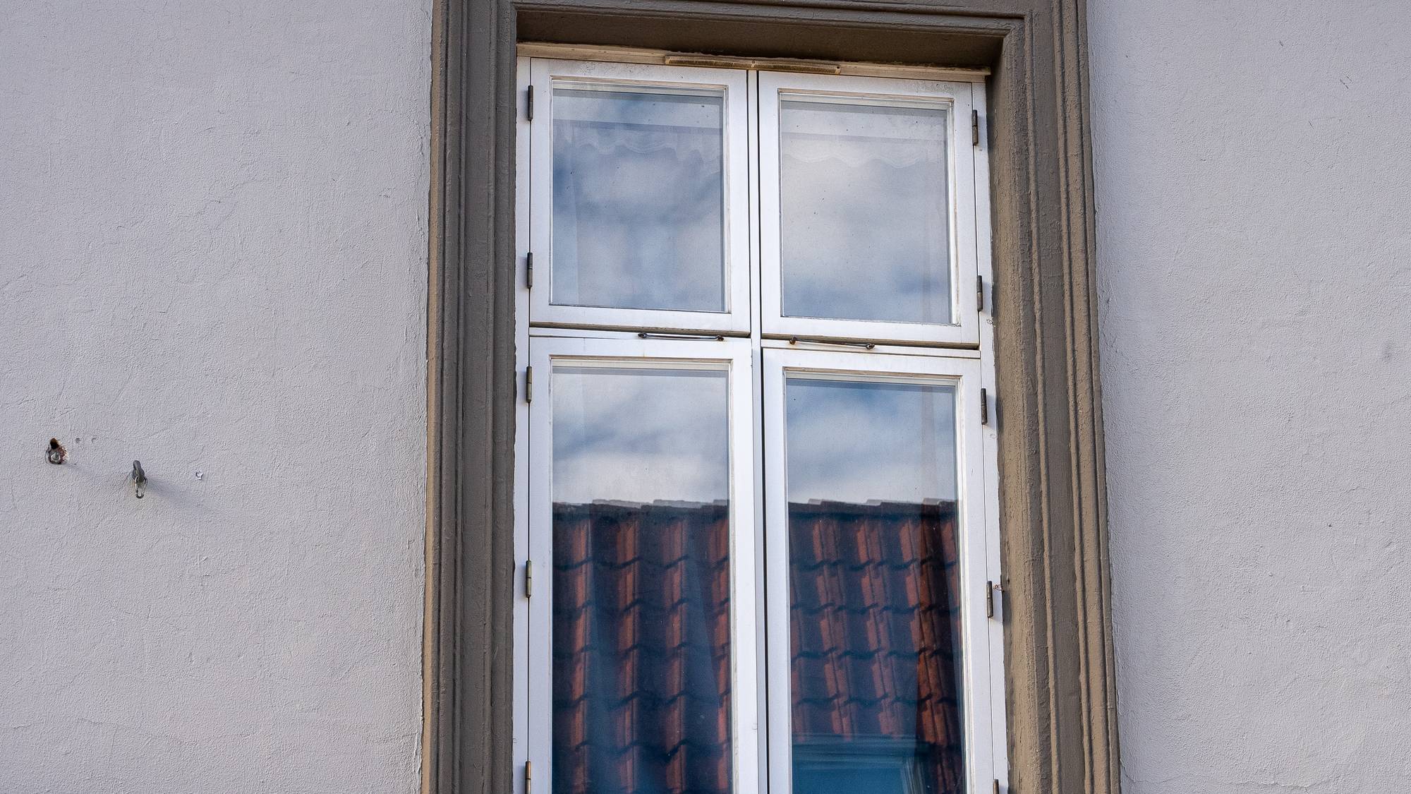 Hvitmalt vindu med fire felt i en lys grå bygningsvegg. Refleksjoner av takstein og himmel i glasset. (Bildebeskrivelsen er laget av en KI-tjeneste)