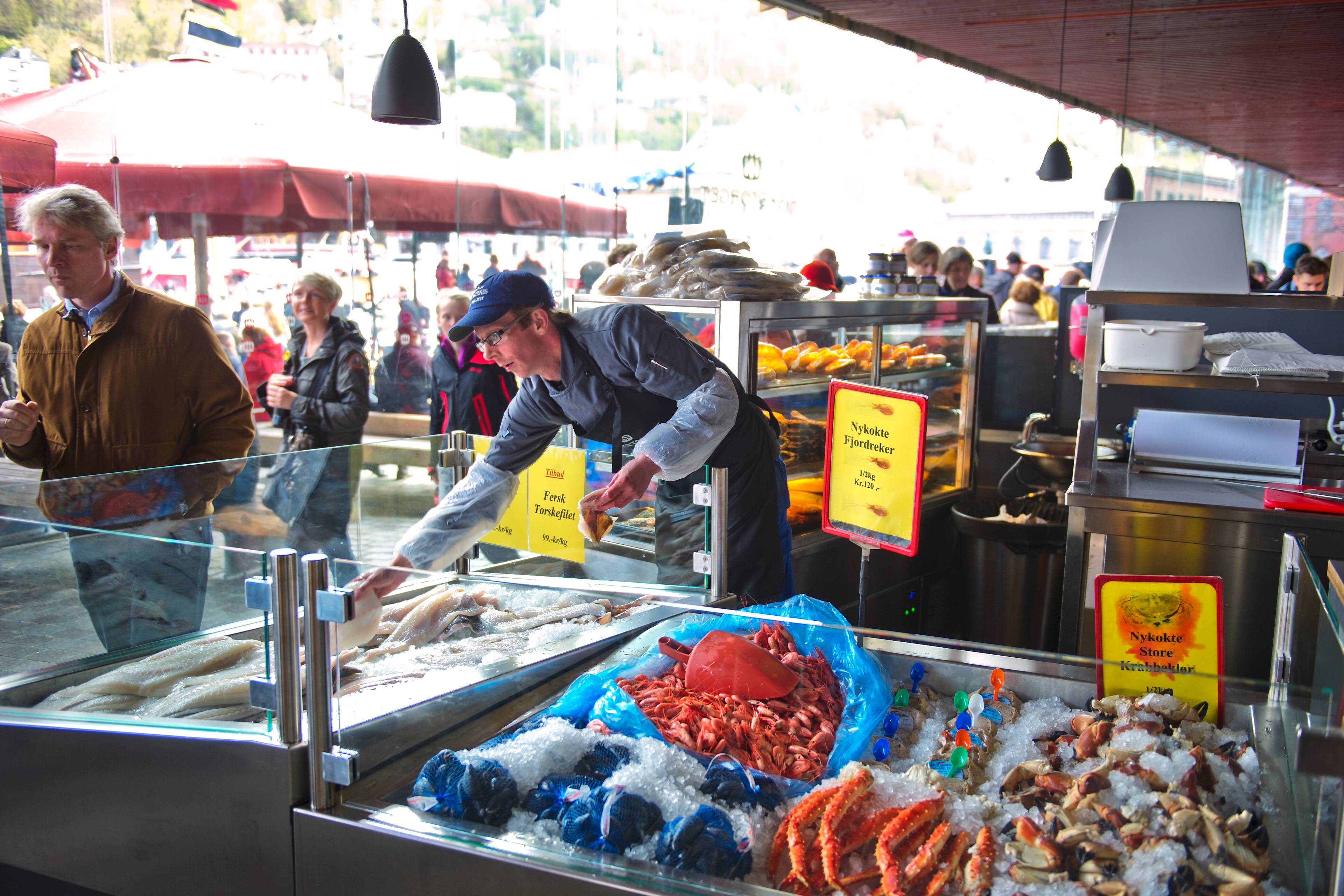 Ekspeditør selger sjømat ved fisketorget i Bergen. Kunder står i bakgrunnen. (Bildebeskrivelsen er laget av en KI-tjeneste)