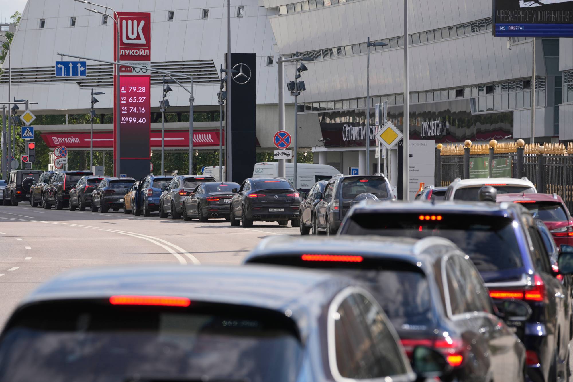 Trafikksituasjon med biler i kø ved en bensinstasjon og en stor kontorbygning. (Bildebeskrivelsen er laget av en KI-tjeneste)