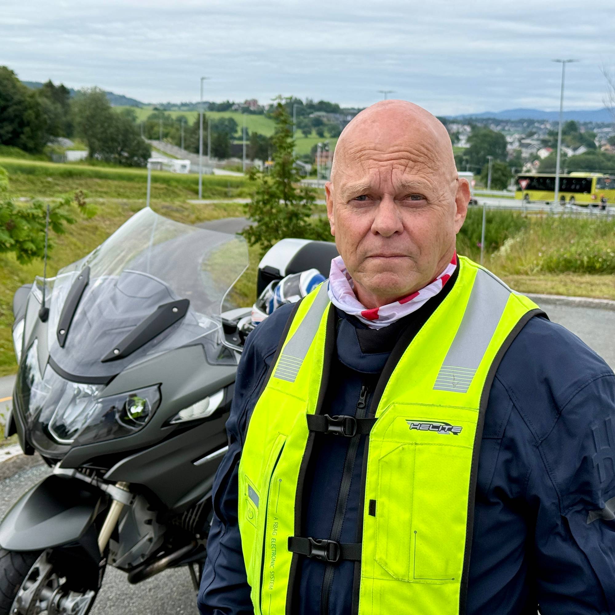 Mann i mc-jakke med gul refleksvest står ved siden av en motorsykkel utendørs.