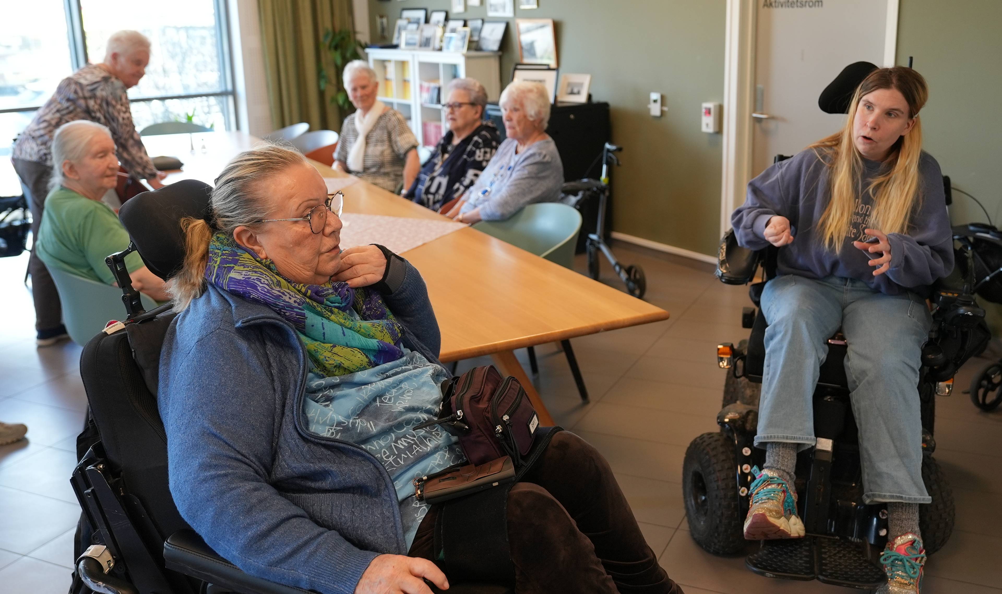 Eldre kvinner pluss to kvinner i rullestol rundt et bord.