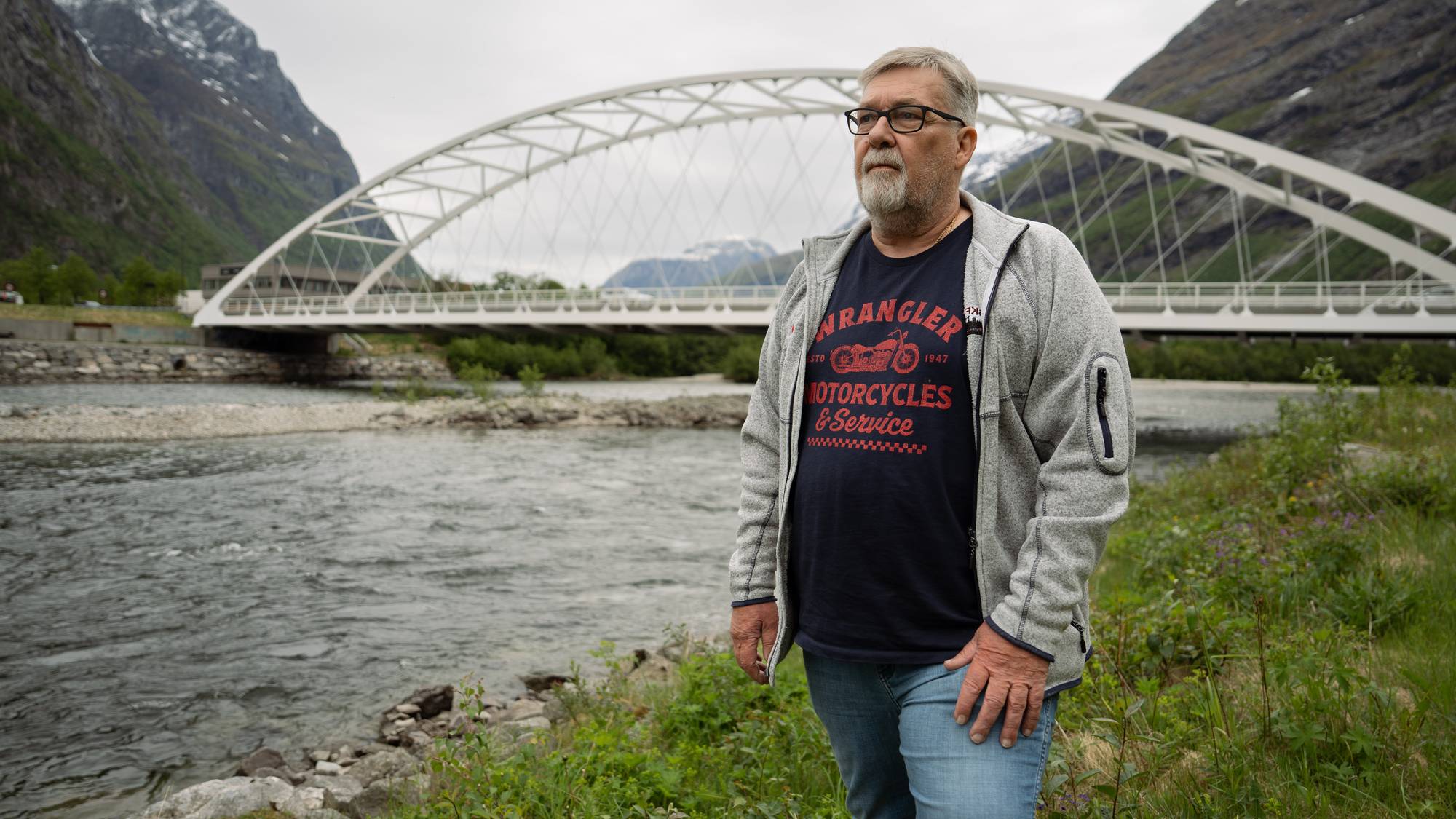 En mann i 60-årene står foran en elv og en hvit buebro. Han har på seg en grå fleecejakke over en mørkeblå t-skjorte med tekst og bilde av en motorsykkel. I bakgrunnen sees fjellsider med snø på toppene under en overskyet himmel. (Bildebeskrivelsen er laget av en KI-tjeneste)