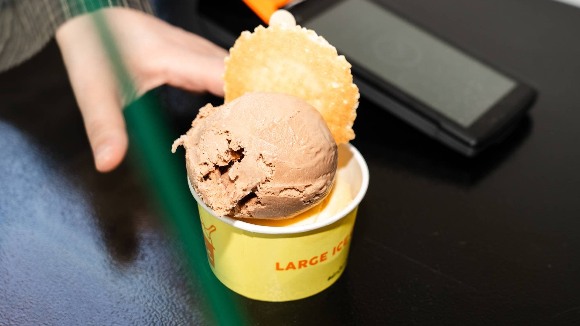 En hånd rekker mot en kopp med to kuler sjokoladeis. En kjeks er stukket inn i isen. Koppen har teksten "LARGE ICE" trykt på siden. I bakgrunnen ligger en mobiltelefon og en oransje gjenstand. 